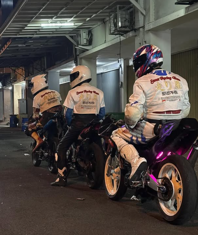 Three motorcycle racers in racing suits and helmets sitting on their bikes in a garage or pit area.