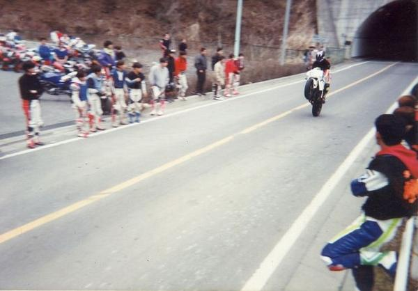 A motorcycle racing out of a tunnel on a mountain road, with spectators of various ages watching from the roadside, some sitting and some standing.