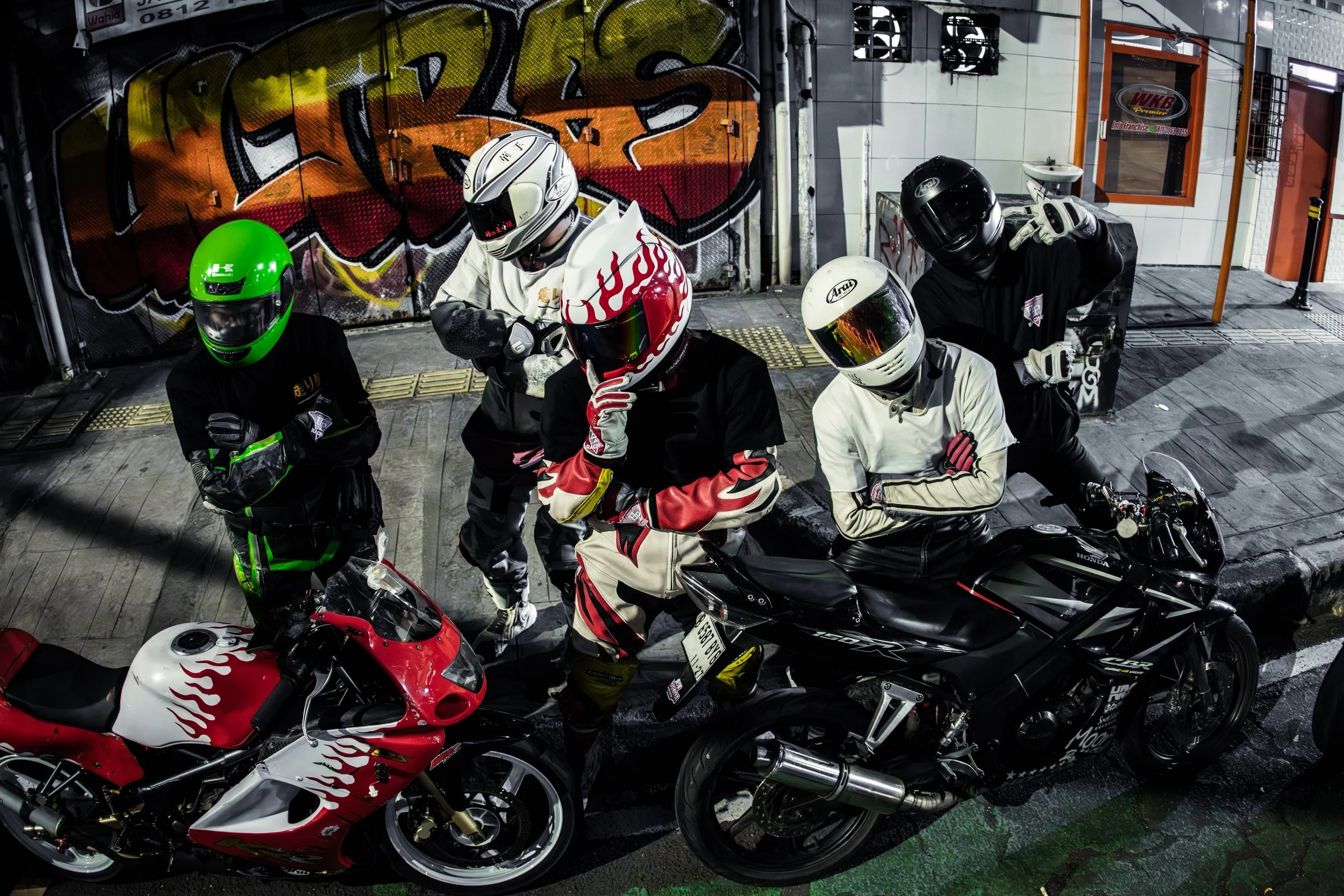 Group of four Hashiriya wearing helmets and protective gear, gathered in front of a graffiti-covered wall, with their motorcycles parked nearby.