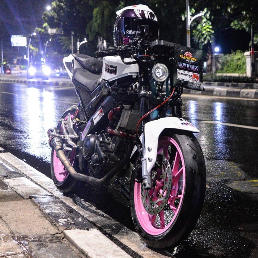 A custom motorcycle parked on a wet street at night, featuring pink rims, a black and white body, and various stickers and decals on the front. It has a helmet resting on the handlebars.