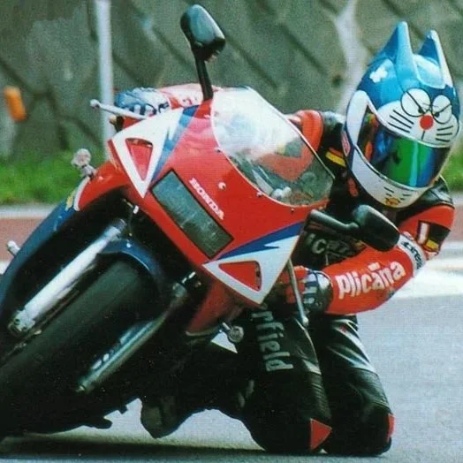 A person riding a red, white, and blue Honda motorcycle with a Doraemon helmet, leaning into a turn on a paved road with green trees in the background.
