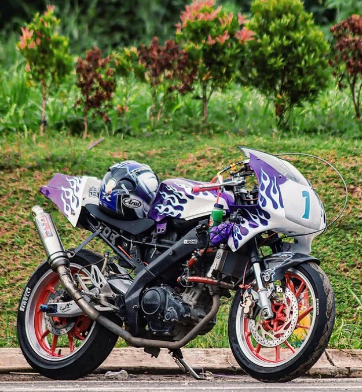A customized purple and white racing motorcycle with flame decals, a helmet on the seat, parked on a roadside in front of green foliage.