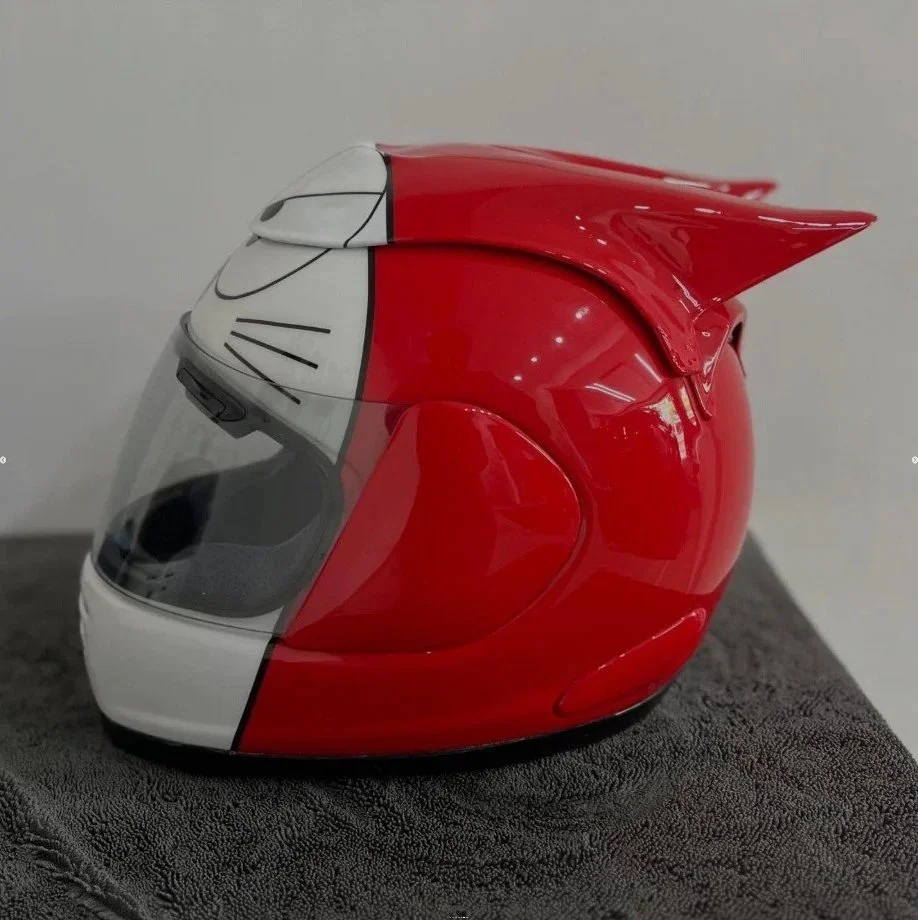 A motorcycle helmet painted to resemble the cartoon character Hello Kitty, with a white face, black whiskers, and a red body with small white accents.