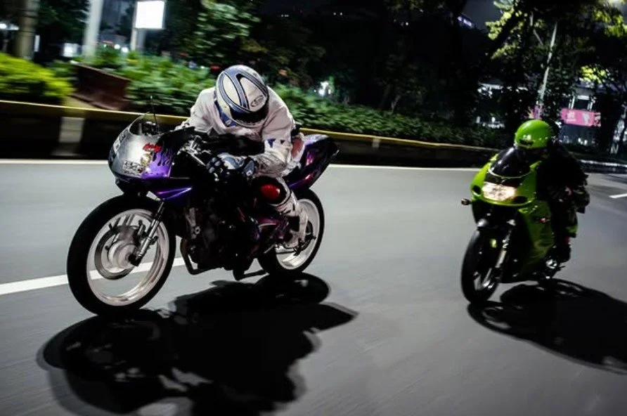 Two motorcyclists riding on a city street at night, one in purple gear and the other in green gear.