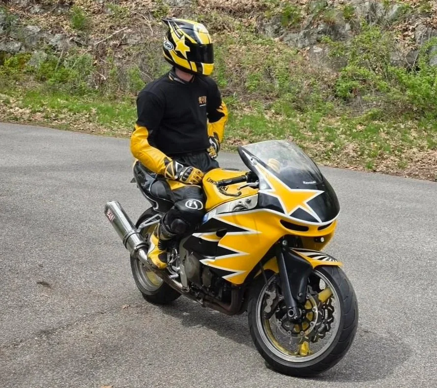 Motorcyclist wearing a black helmet with yellow designs and a black and yellow racing suit, sitting on a yellow and black sports motorcycle on the road with greenery in the background.