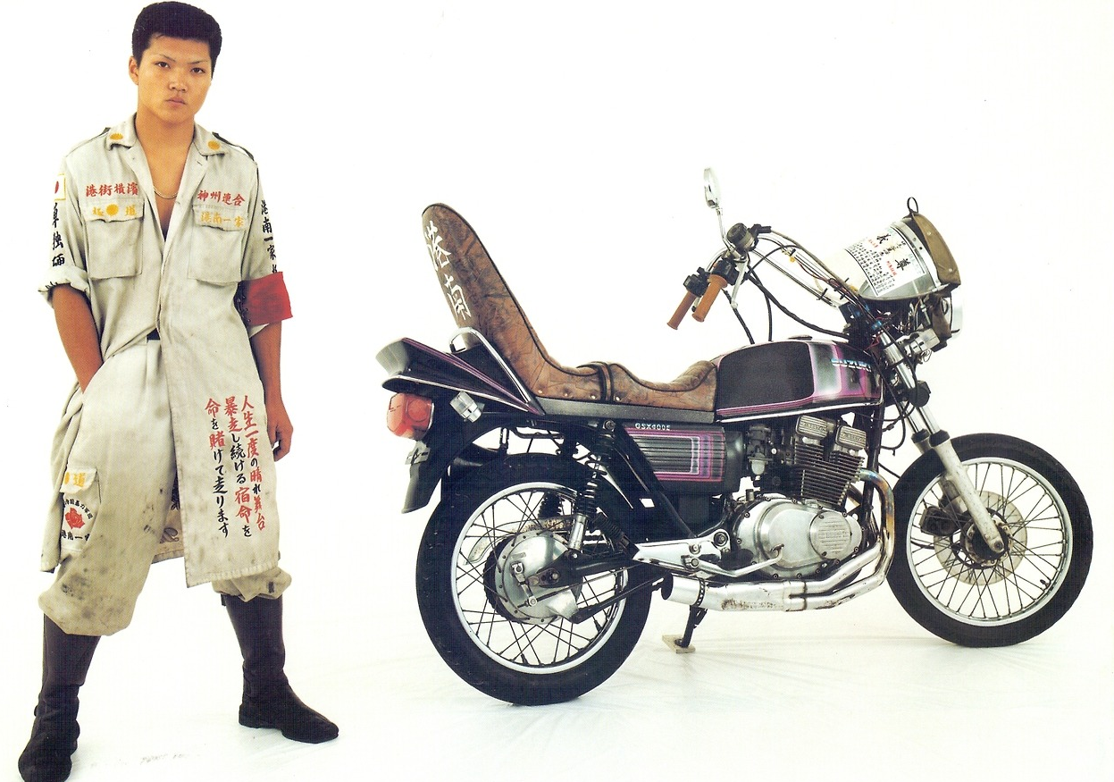 A young man in a beige jumpsuit with Japanese text stands next to a vintage motorcycle with a high seat and Japanese writing, against a plain white background.