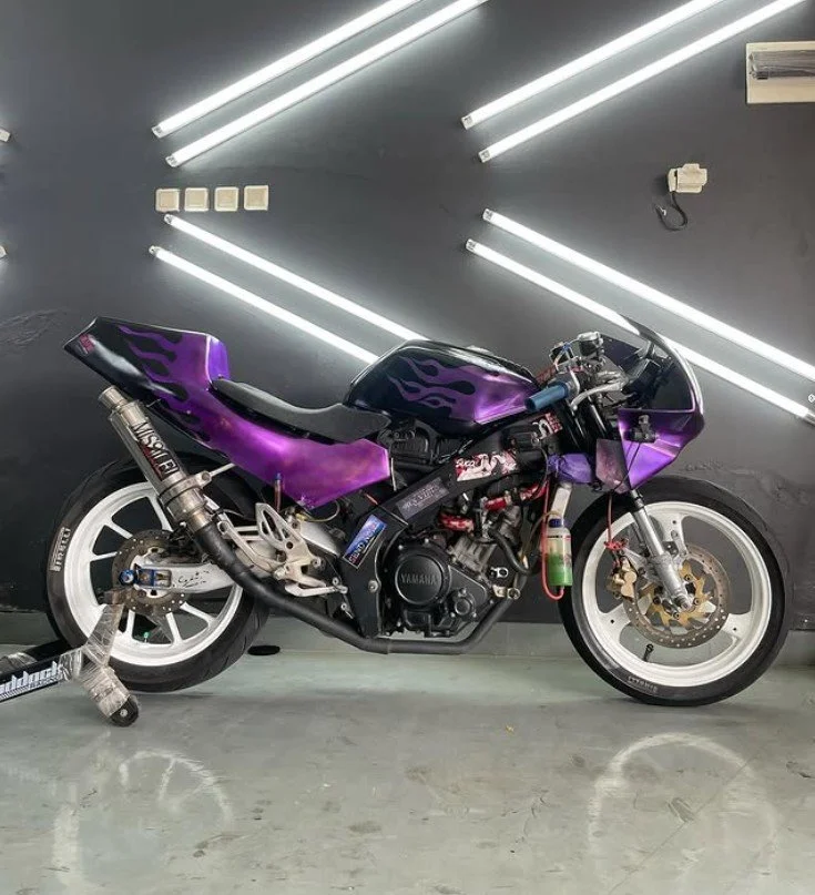A purple motorcycle with flames on the fairings, white wheels, and aftermarket exhaust, parked indoors against a dark wall with LED light strips.