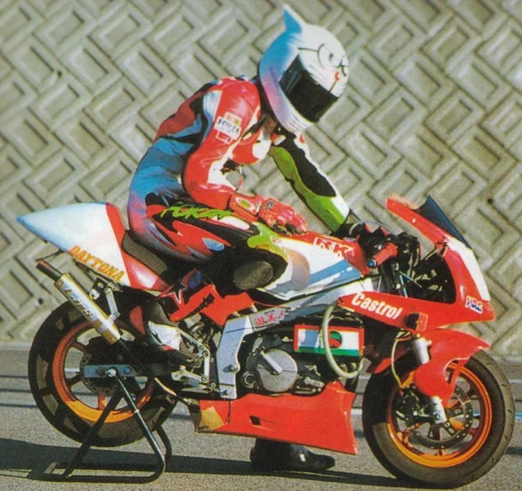 Motorsport racer in racing suit and helmet riding a red racing motorcycle with sponsor logos, against a textured background.