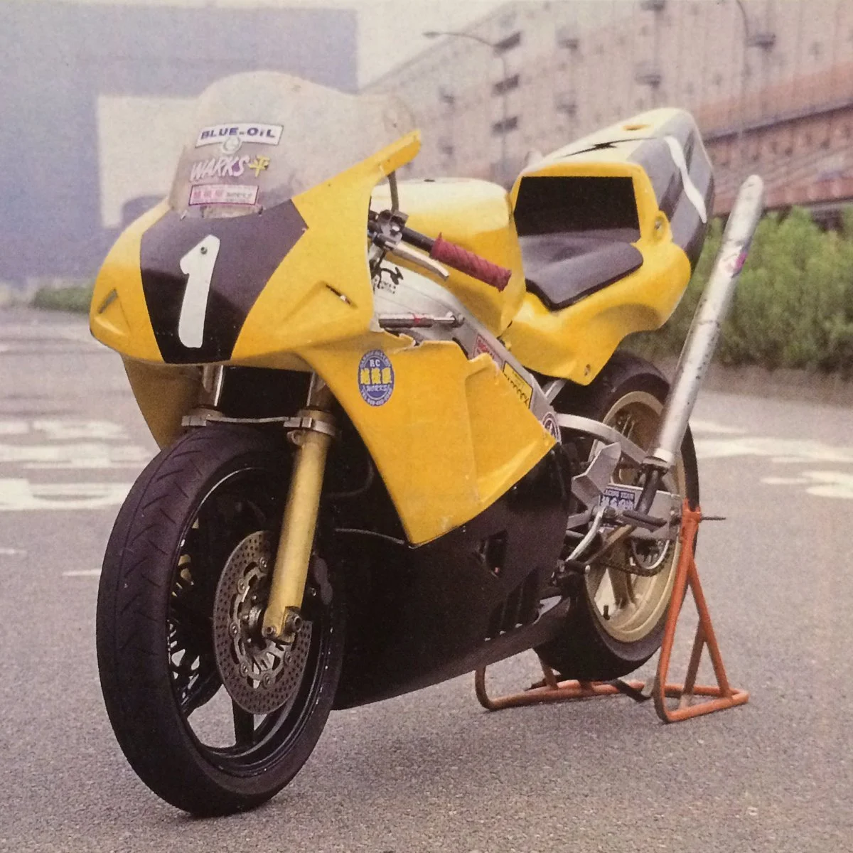 Yellow racing motorcycle with race number 1 and sponsorship stickers, resting on a stand in an urban parking lot.