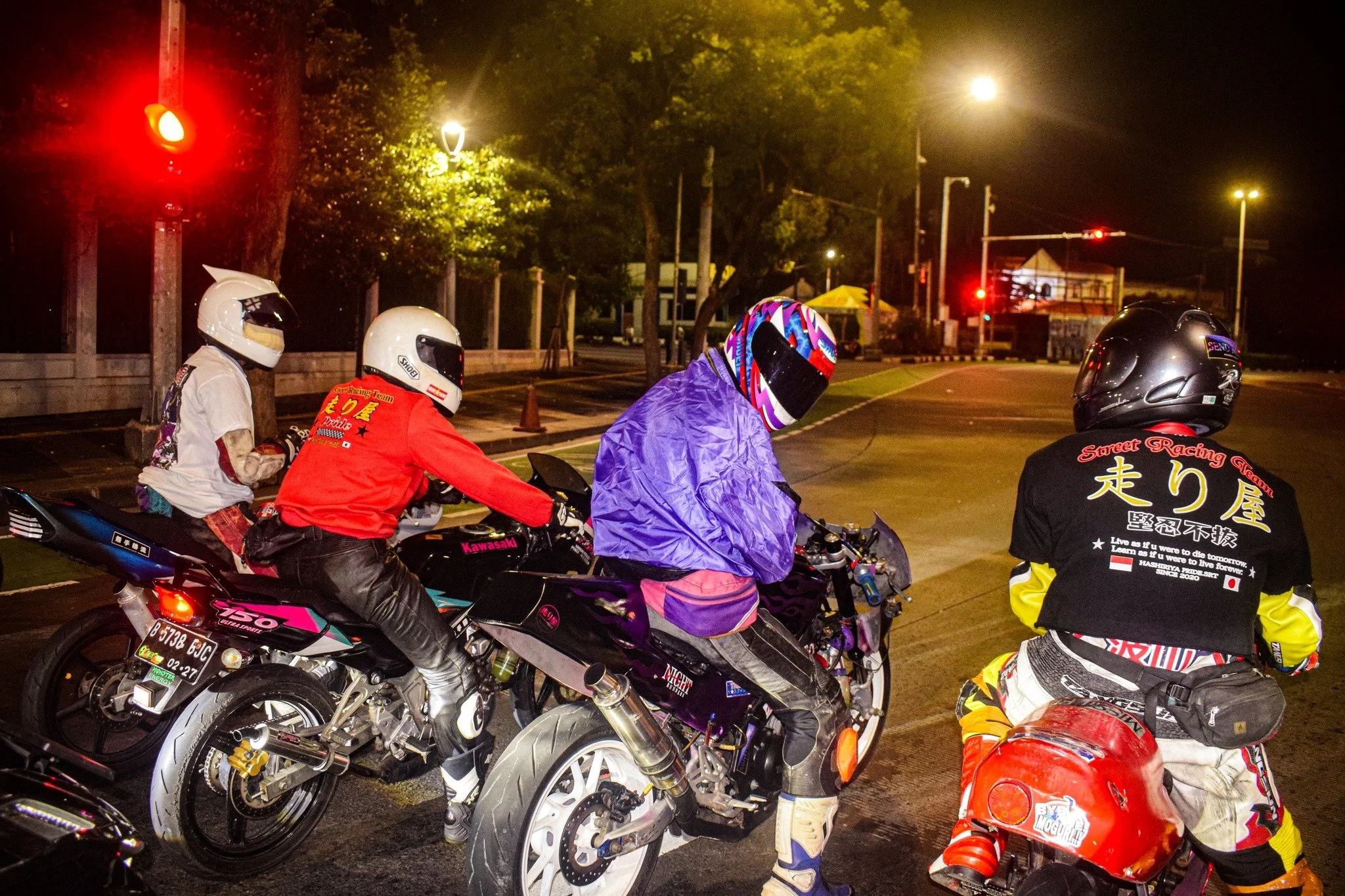 Group of motorcyclists in racing gear and helmets gathered on a city street at night.