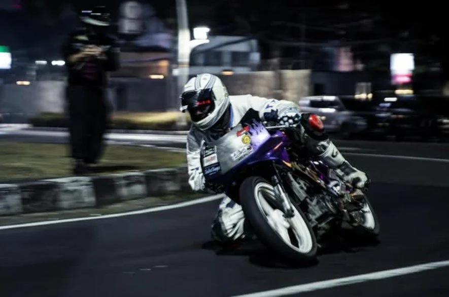 Motorcycle racing at night, rider leaning sharply into a turn on a city street.