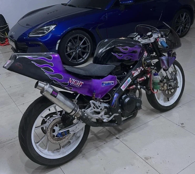 A motorcycle with a purple and black paint scheme, flames on the rear fender, and the words "Night Runnin'" parked next to a blue sports car in a garage.