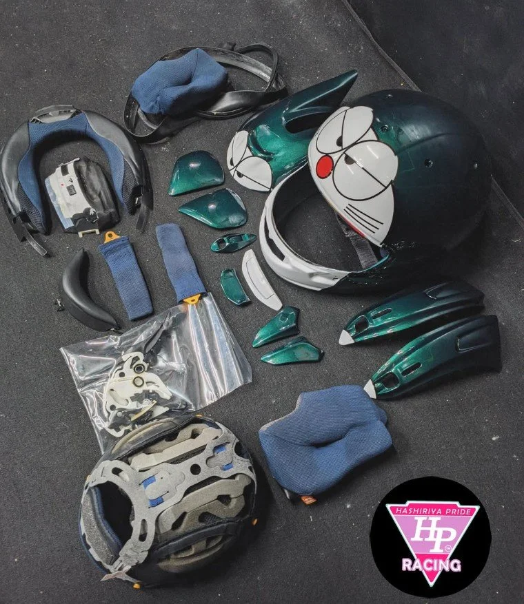 Disassembled green and white Doraemon helmet, motorcycle gloves, and other protective gear on a black surface, with a logo reading 'Hashiriya Pride Racing' in the lower right corner.