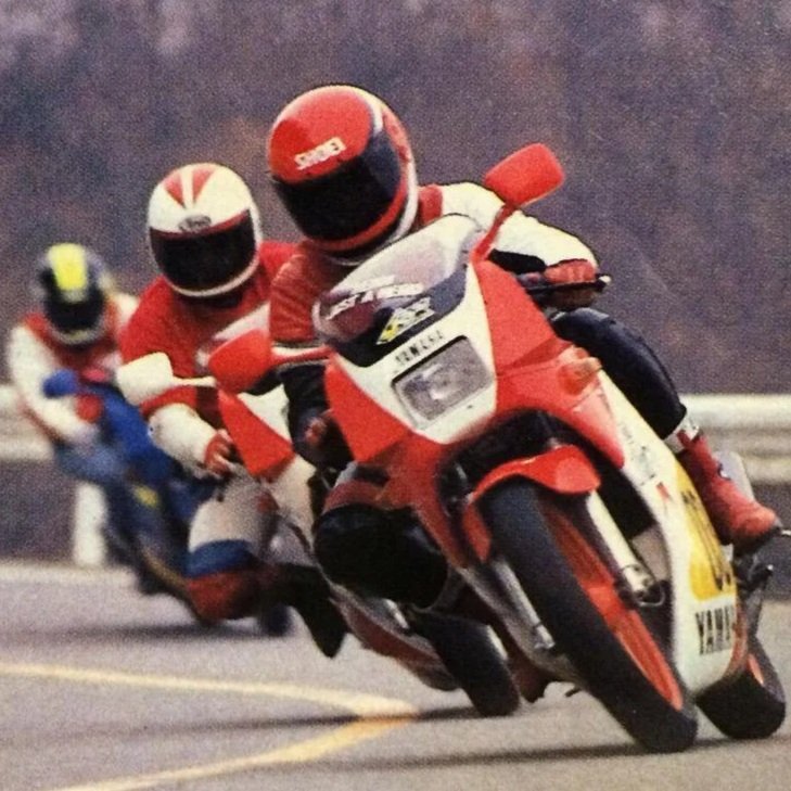 Three motorcyclists racing on a track, leaning into a turn with the lead rider on a red and white motorcycle.