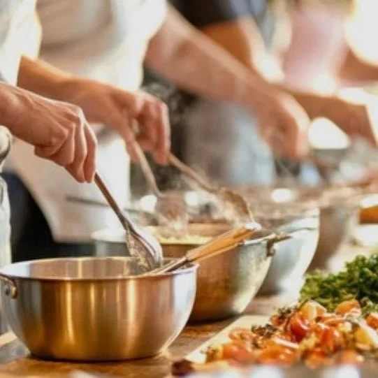 People preparing food at a buffet or cooking station, using tongs to serve dishes into bowls.