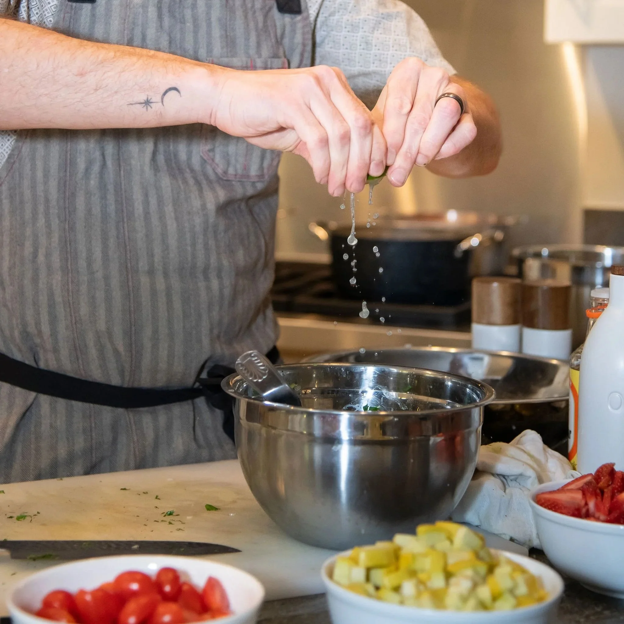 People preparing food in a kitchen with bowls of cherry tomatoes, diced yellow vegetables, and chopped herbs on the countertop, and hands washing or rinsing vegetables.