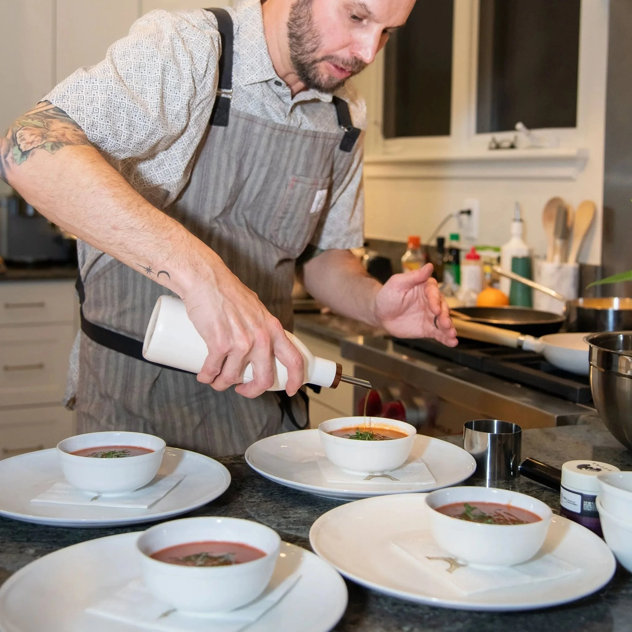 A man with tattoos on his arms and a beard, wearing a patterned shirt and striped apron, preparing food in a kitchen. Several bowls of soup are on the counter in front of him, and he is adding ingredients to them.