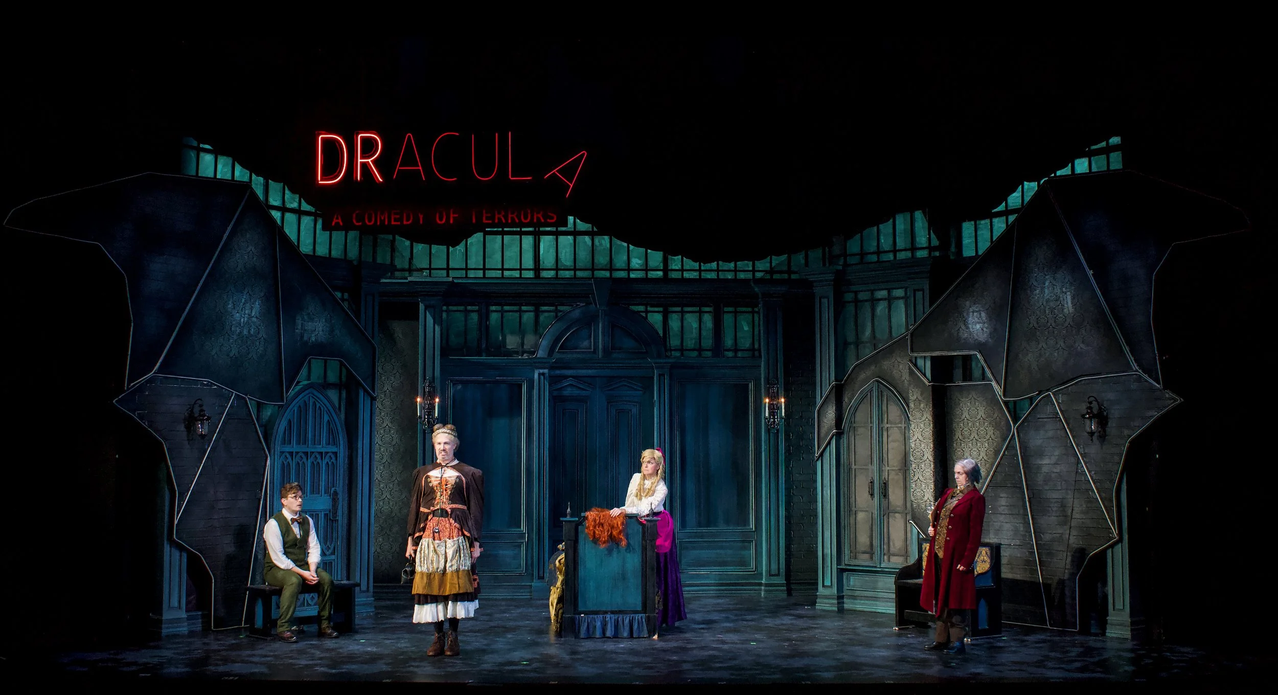 Scene from a theatrical play titled 'Dracula: A Comedy of Terrohs' featuring actors in period costumes standing and sitting on stage in front of a gothic-style set with large windows and dark blue doors.