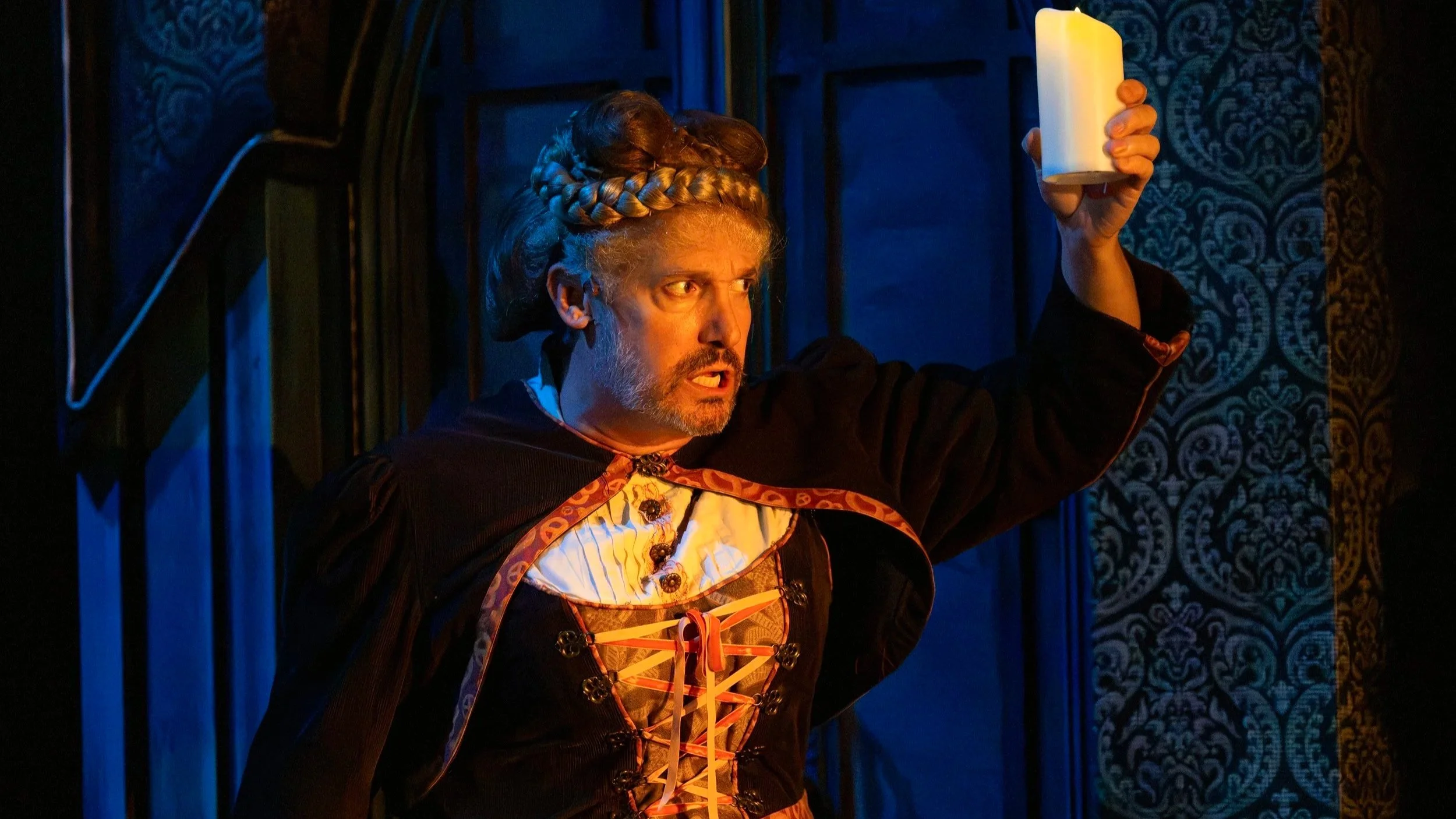 A person dressed in a witch or wizard costume holding a large white candle, with a dark background and decorative blue patterned walls.