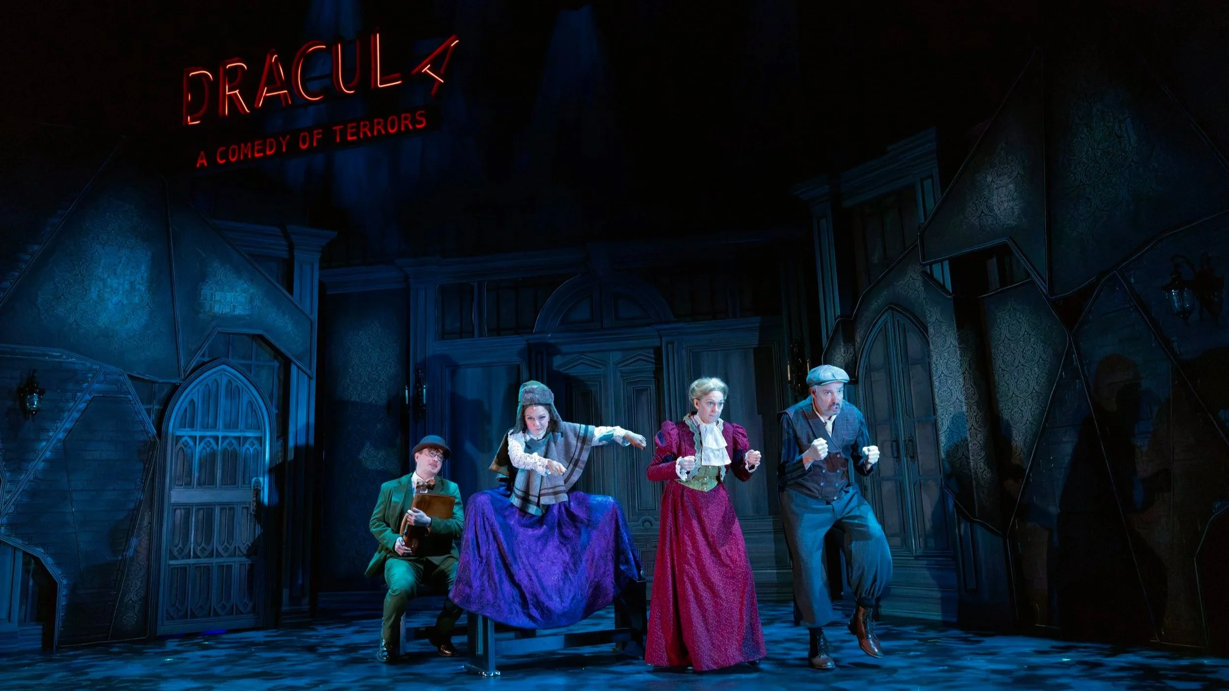 Actors performing on stage in a theatrical production, with a backdrop featuring gothic architecture design and a neon sign that reads 'Dracula: A Comedy of Terrors'.