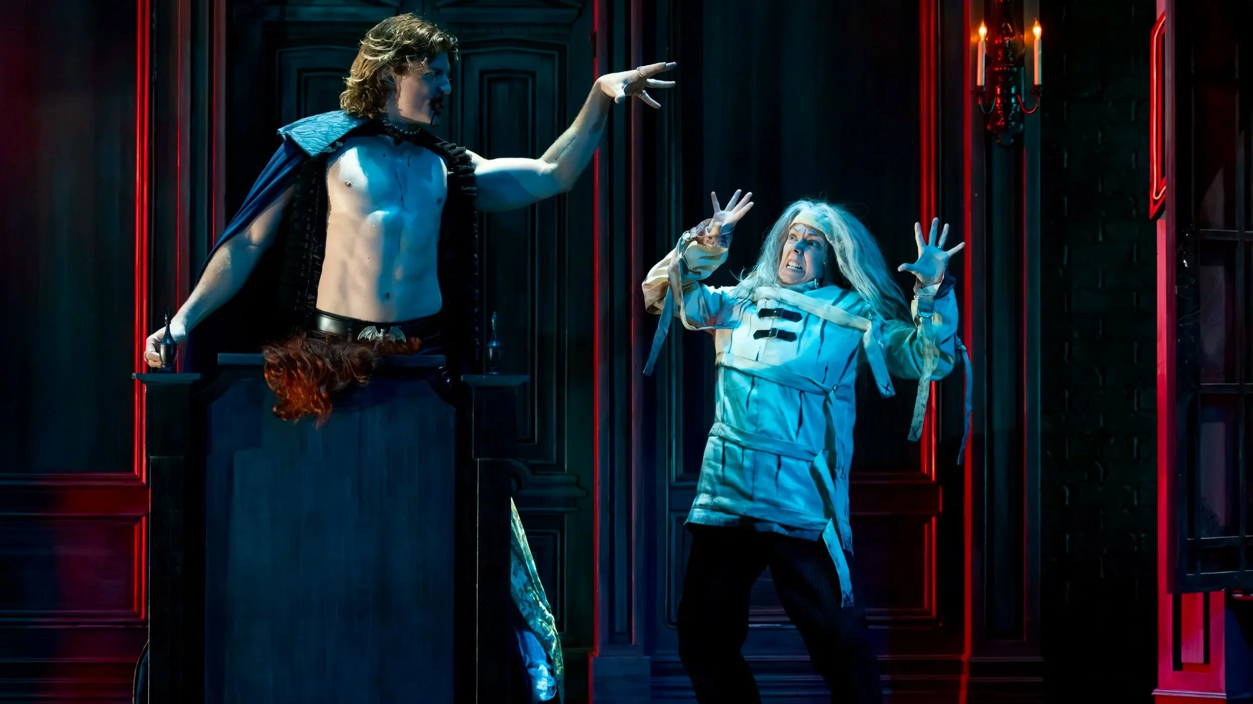 A theatrical scene with a shirtless man dressed as a vampire and a woman in hospital pajamas, both appearing distressed.