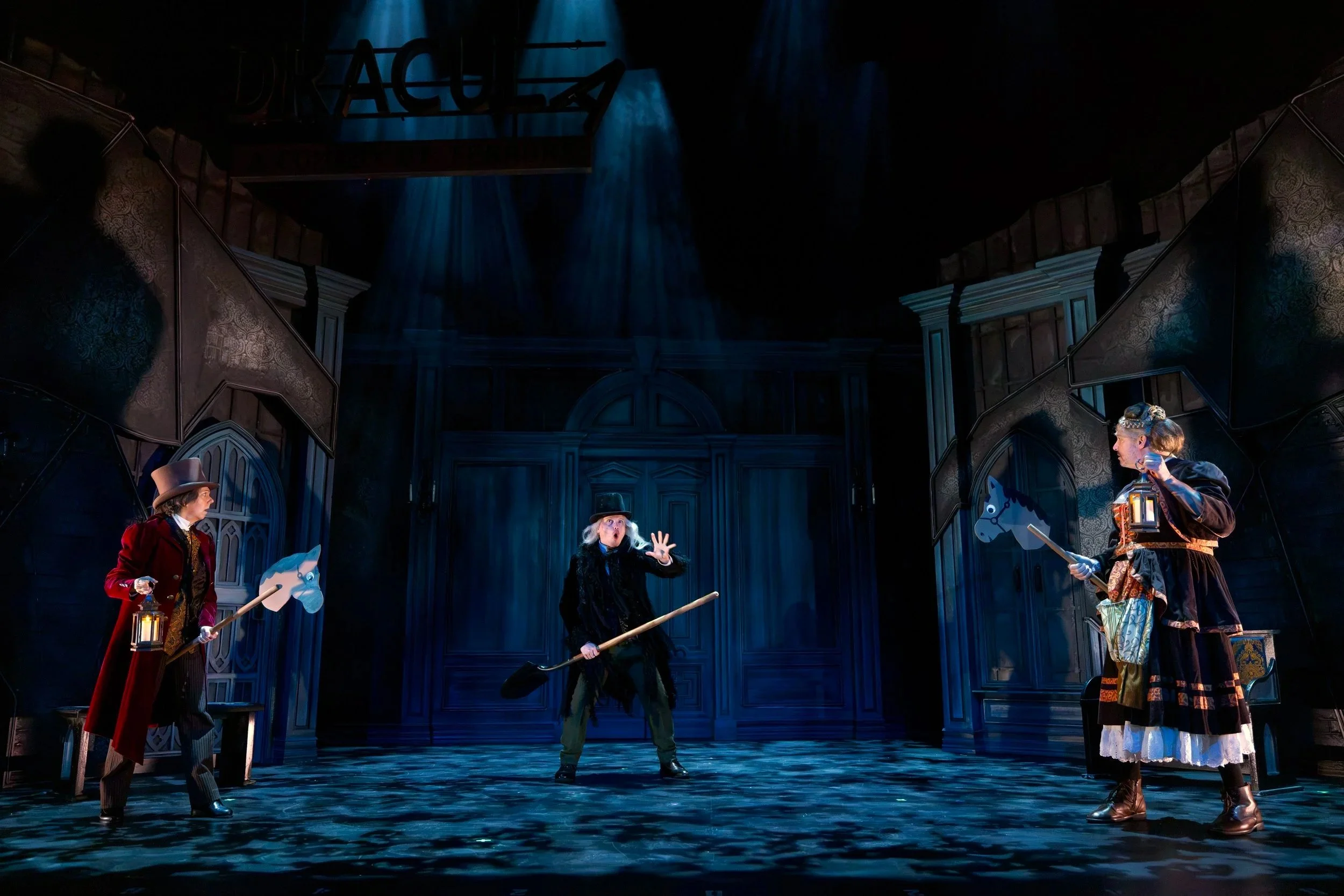The image shows a scene from the musical or play 'Beauty and the Beast' with four characters dressed in period costumes. They are standing on a stage with dark, ornate wooden architectural elements and a sign that partially reads 'DRA...'. The charac
