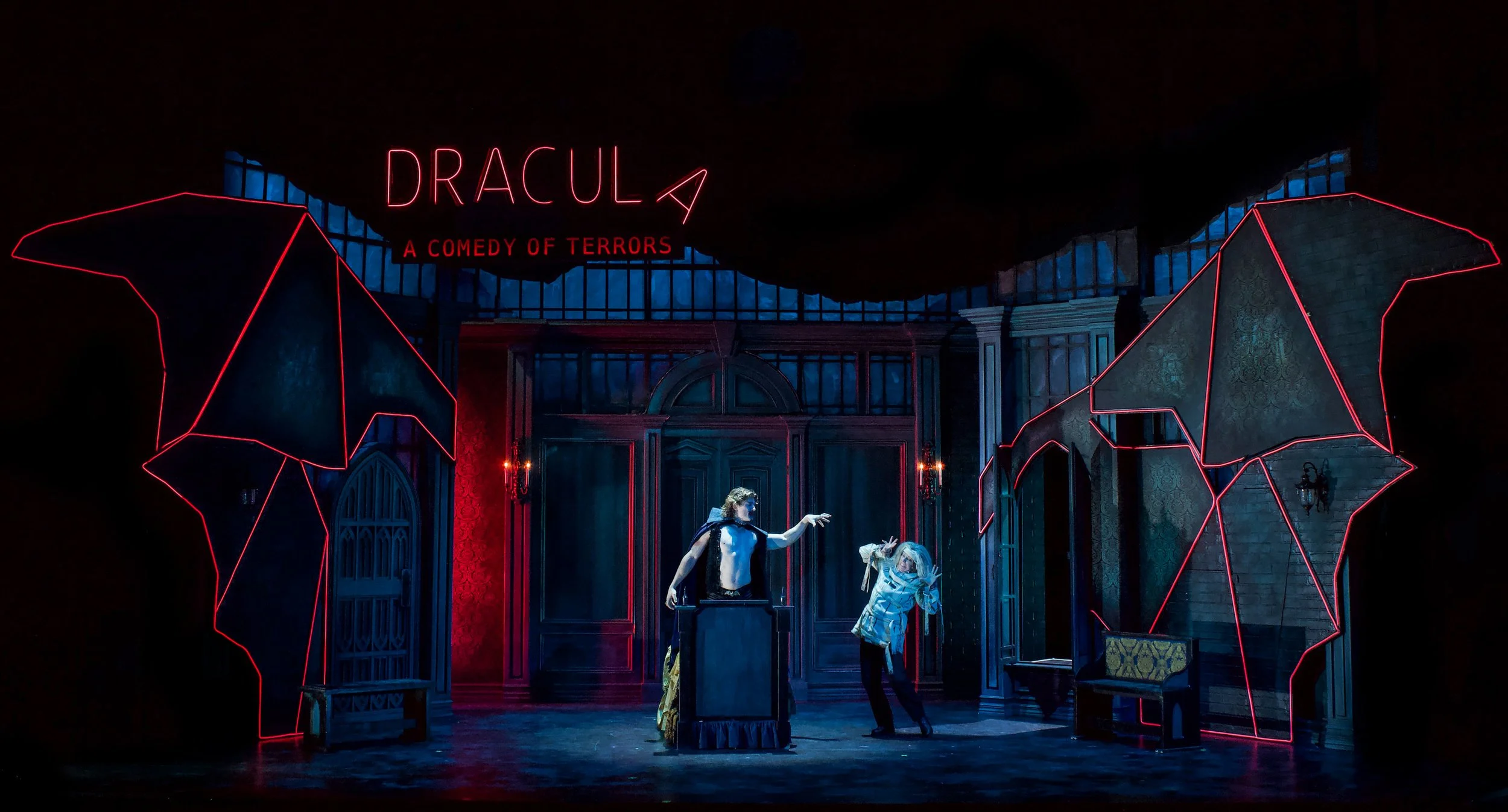 Stage with neon sign reading 'DRACULA' and 'A COMEDY OF TERRORS,' featuring actors in costumes and dramatic lighting.