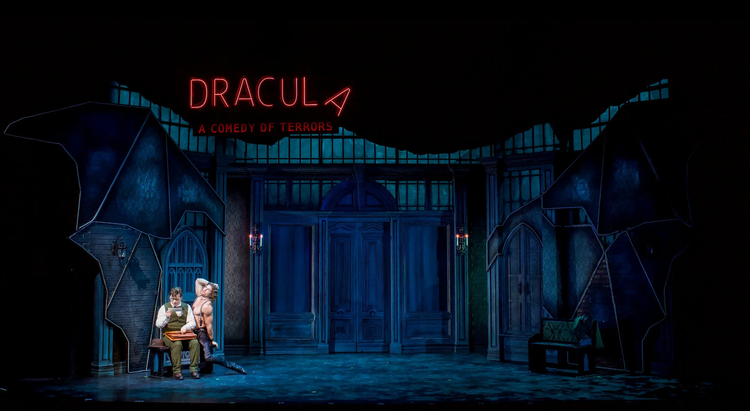 Stage set for a theatrical production of Dracula with gothic architecture and dark lighting, featuring two actors sitting on a bench, one reading a book and the other standing with a hand on their head, and a neon sign that reads 'DRACULA A COMEDY OF