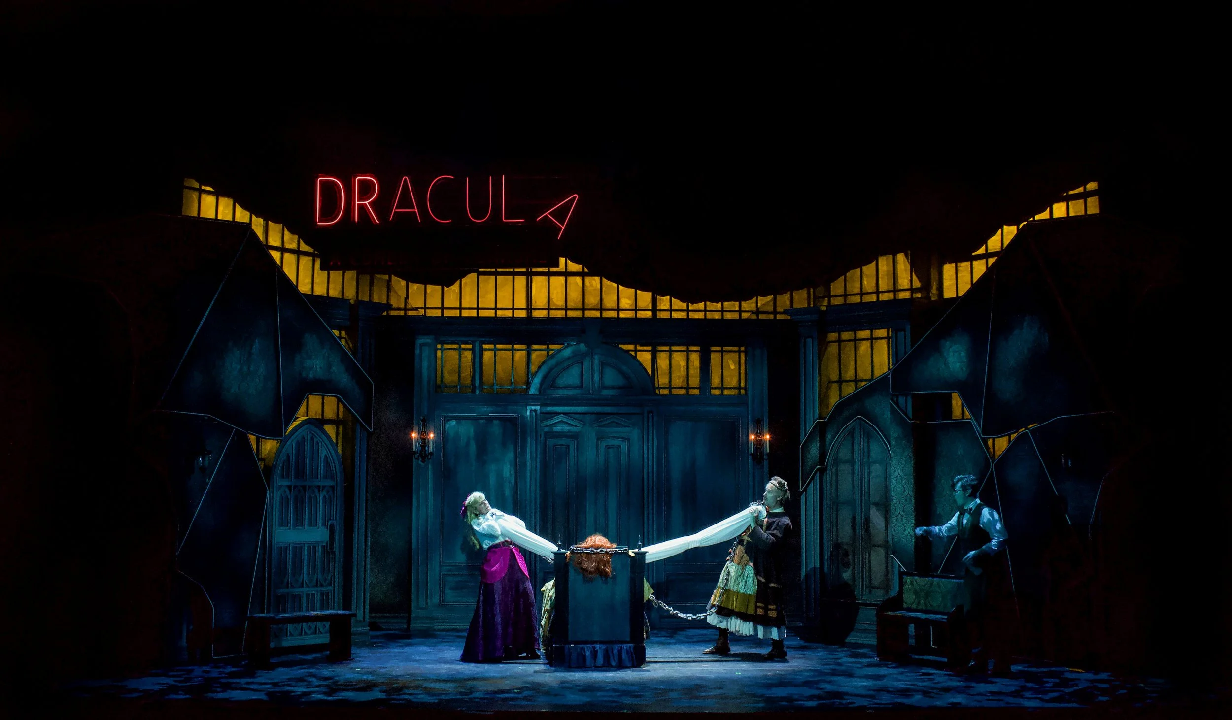 A theatrical stage scene from 'Dracula' with actors in period costumes, a dark Gothic set with large wooden doors, artificial yellow-lit windows, and a neon sign reading 'DRACULA' above the stage.
