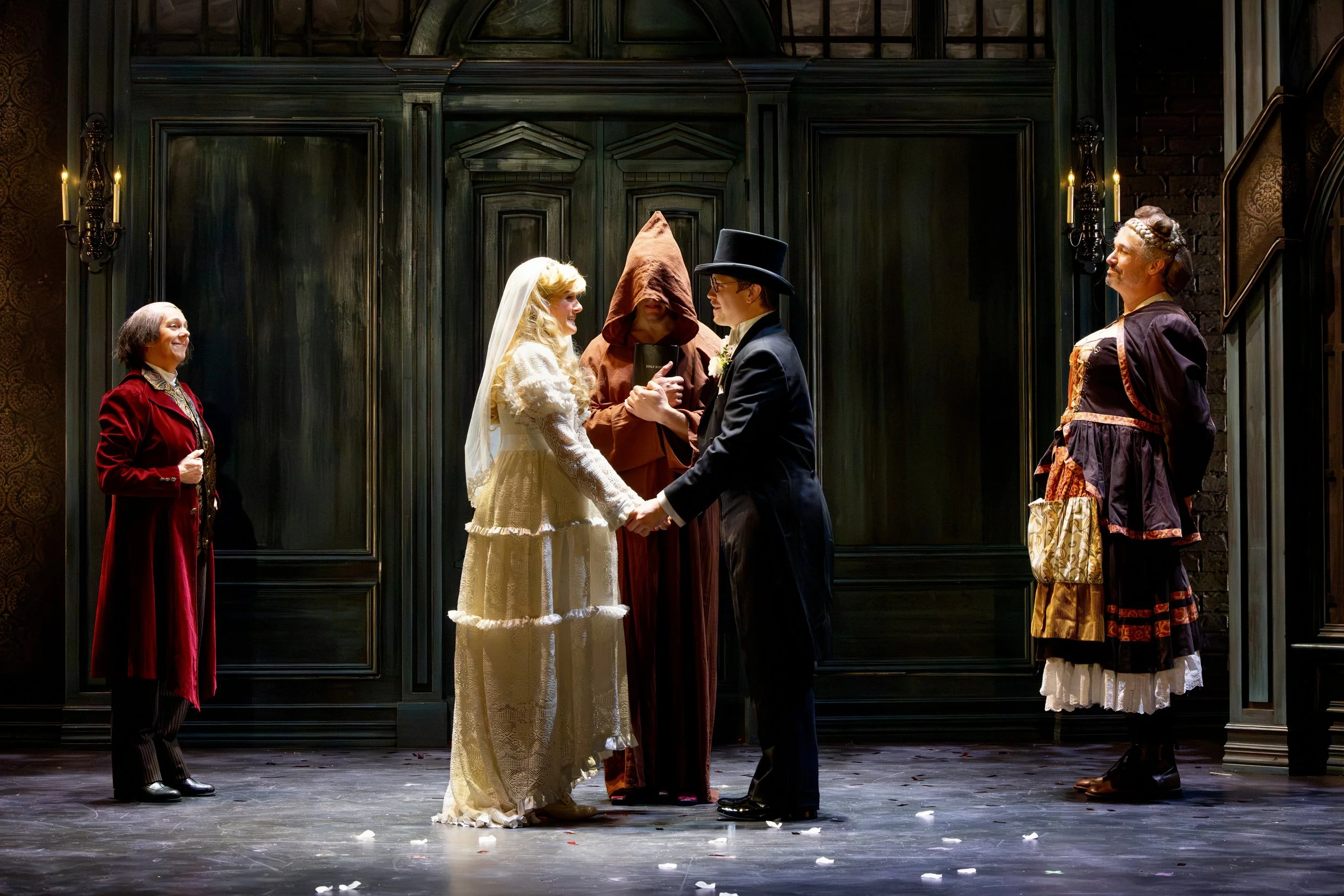 A theatrical stage scene showing a wedding ceremony between a woman in a white vintage wedding dress and veil, and a man in a dark suit and top hat, holding hands and facing each other. Four individuals dressed in vintage or period costumes are stand
