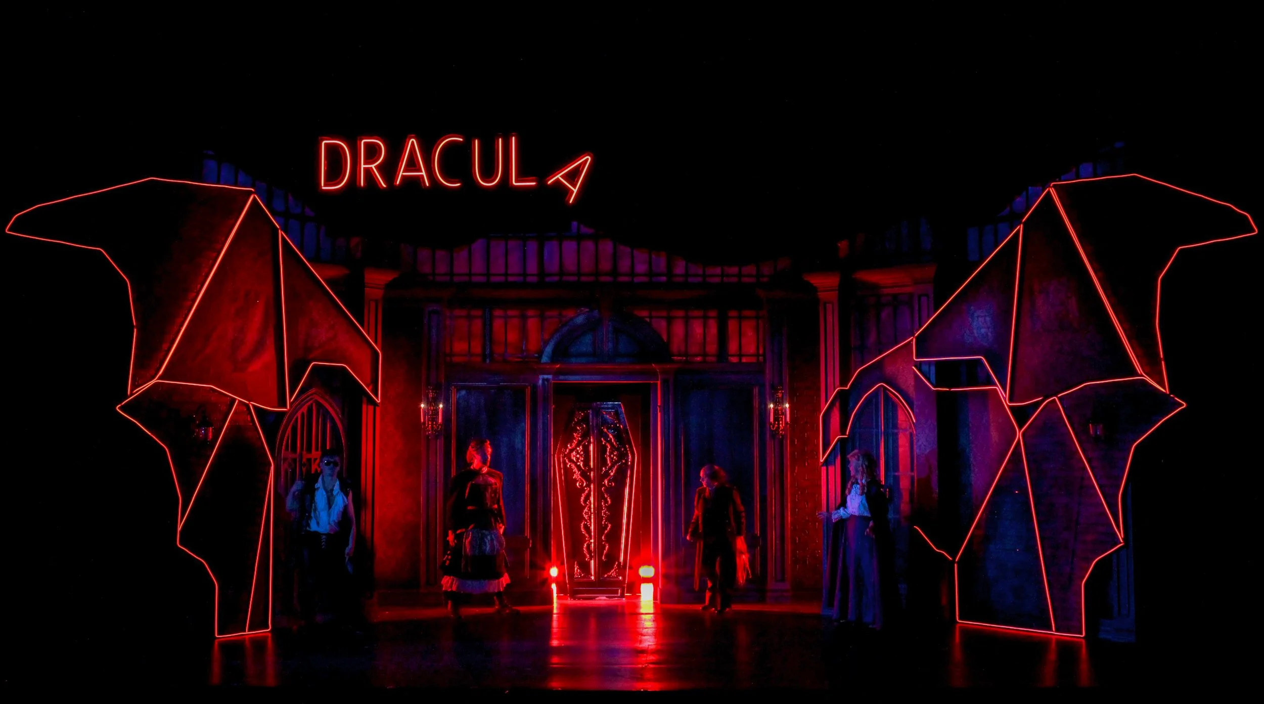 People standing in front of a Gothic-style building decorated with red neon lights, including a sign that reads 'DRACULA'.