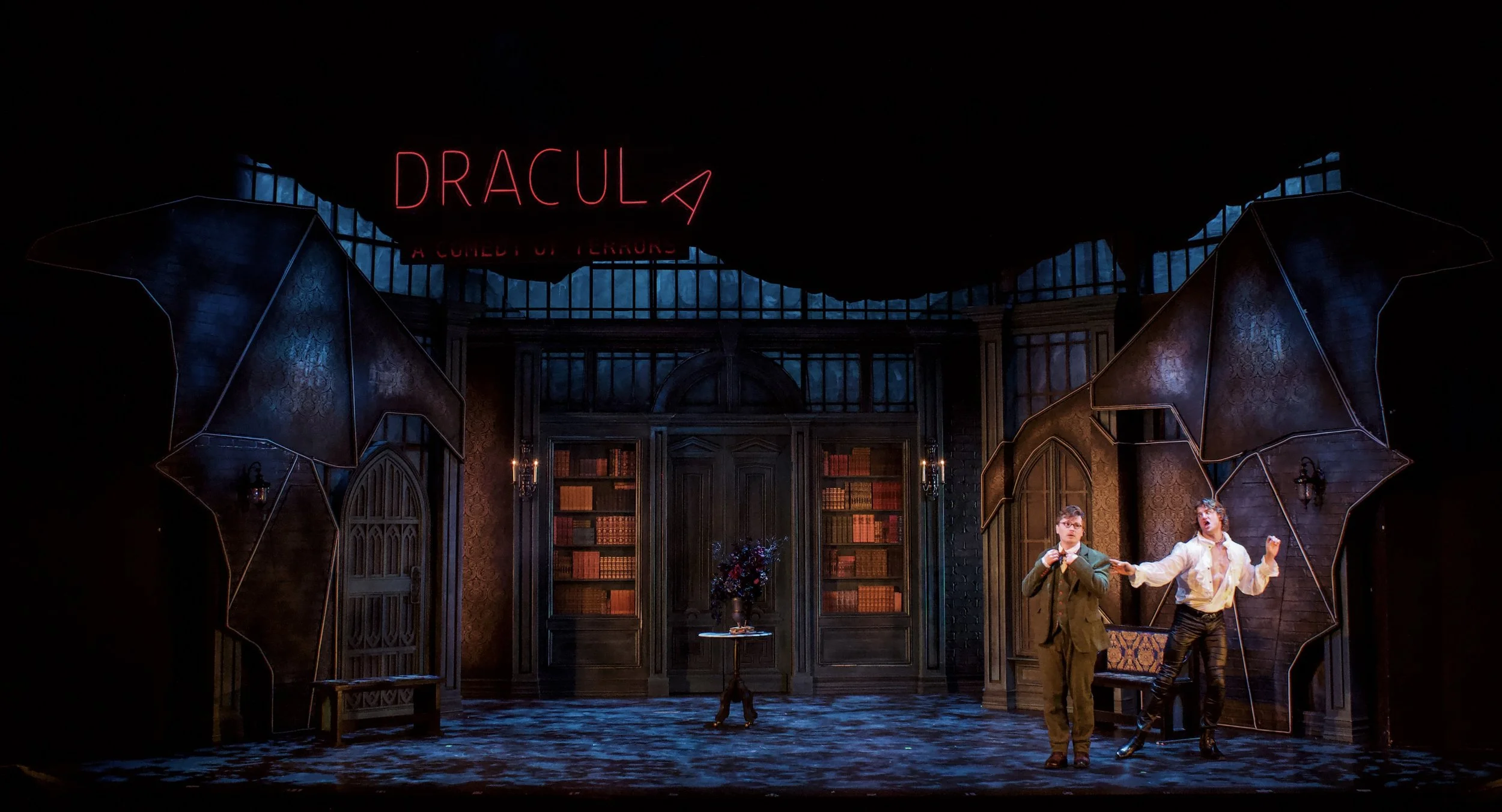 The stage set depicts a gothic library scene with tall bookshelves, dark wood furniture, and ornate decoration. There are two actors in period costumes, one holding a book and the other gesturing dramatically. A neon sign above reads 'DRACULA' with a