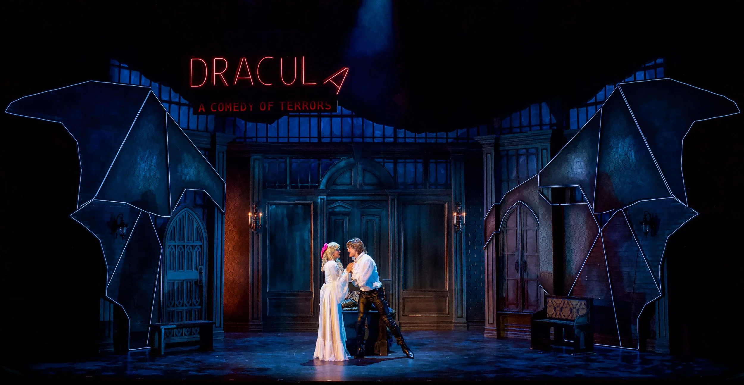 The image shows a scene from a theatrical production of Dracula titled "A Comedy of Terrors." Two actors, dressed in period costume, are standing close together on stage, holding hands and gazing into each other's eyes. The stage has a dark, gothic a