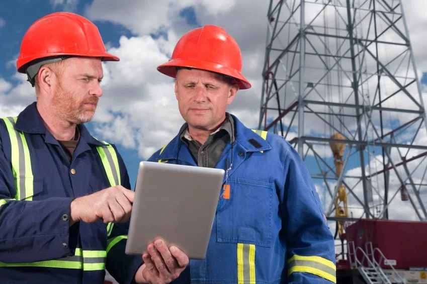 Two male workers in safety helmets and coveralls looking at a tablet in front of a large metal telecommunications tower.