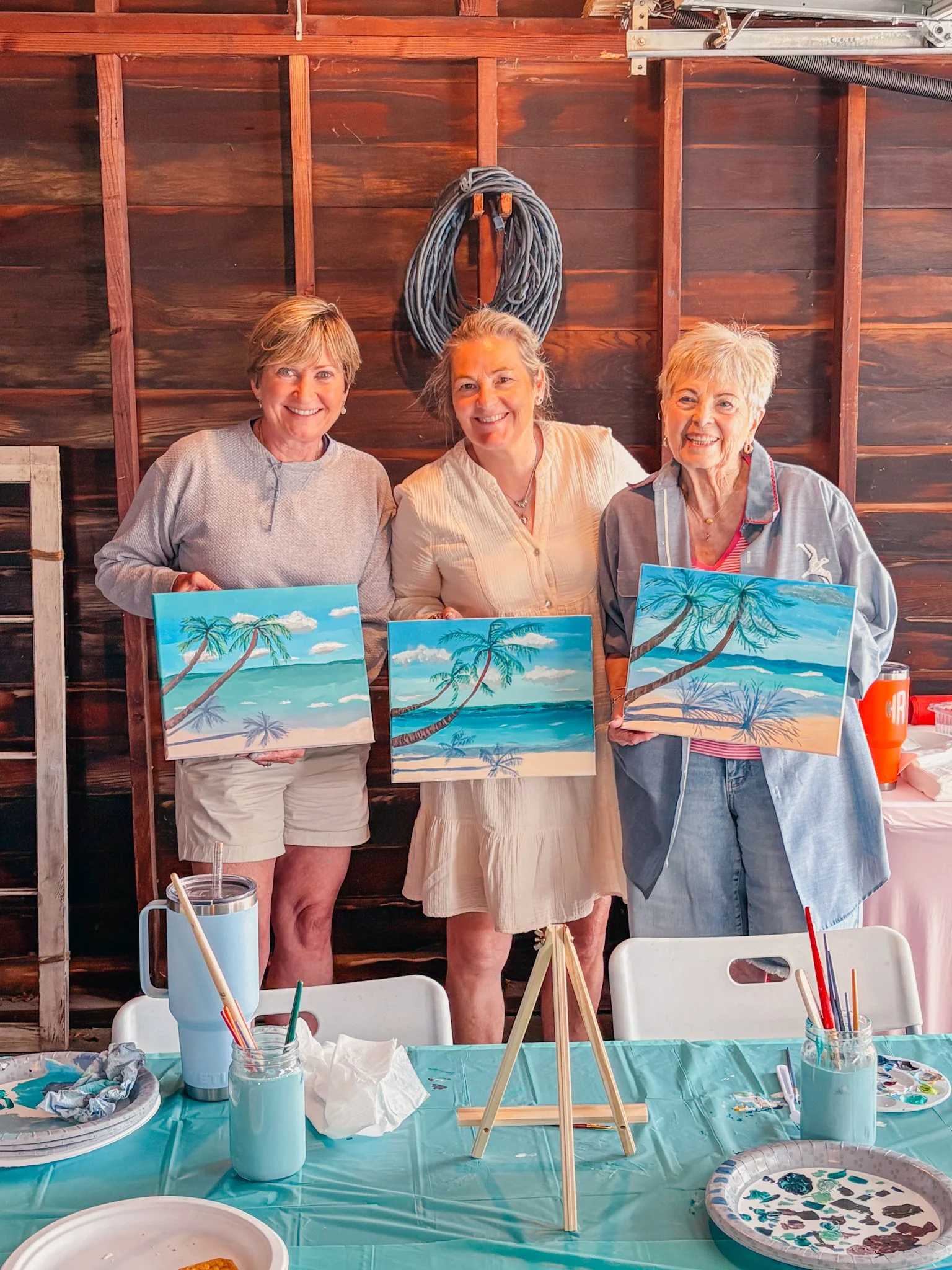 Three women standing behind a table with painting supplies and completed landscape paintings of a beach with palm trees and blue skies. The women are smiling, and the setting appears to be a rustic wooden room or shed.