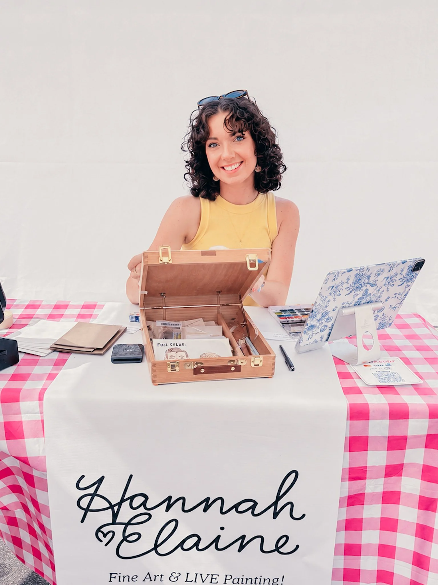A woman sitting at a table covered with a pink and white checkered tablecloth, smiling at the camera with sunglasses on her head. The table has various art supplies, including a wooden case with art materials, a tablet with a floral cover, and other art-related items. A large sign on the front of the table displays the words "Yhamah Elaine" and mentions she offers fine art and live painting.