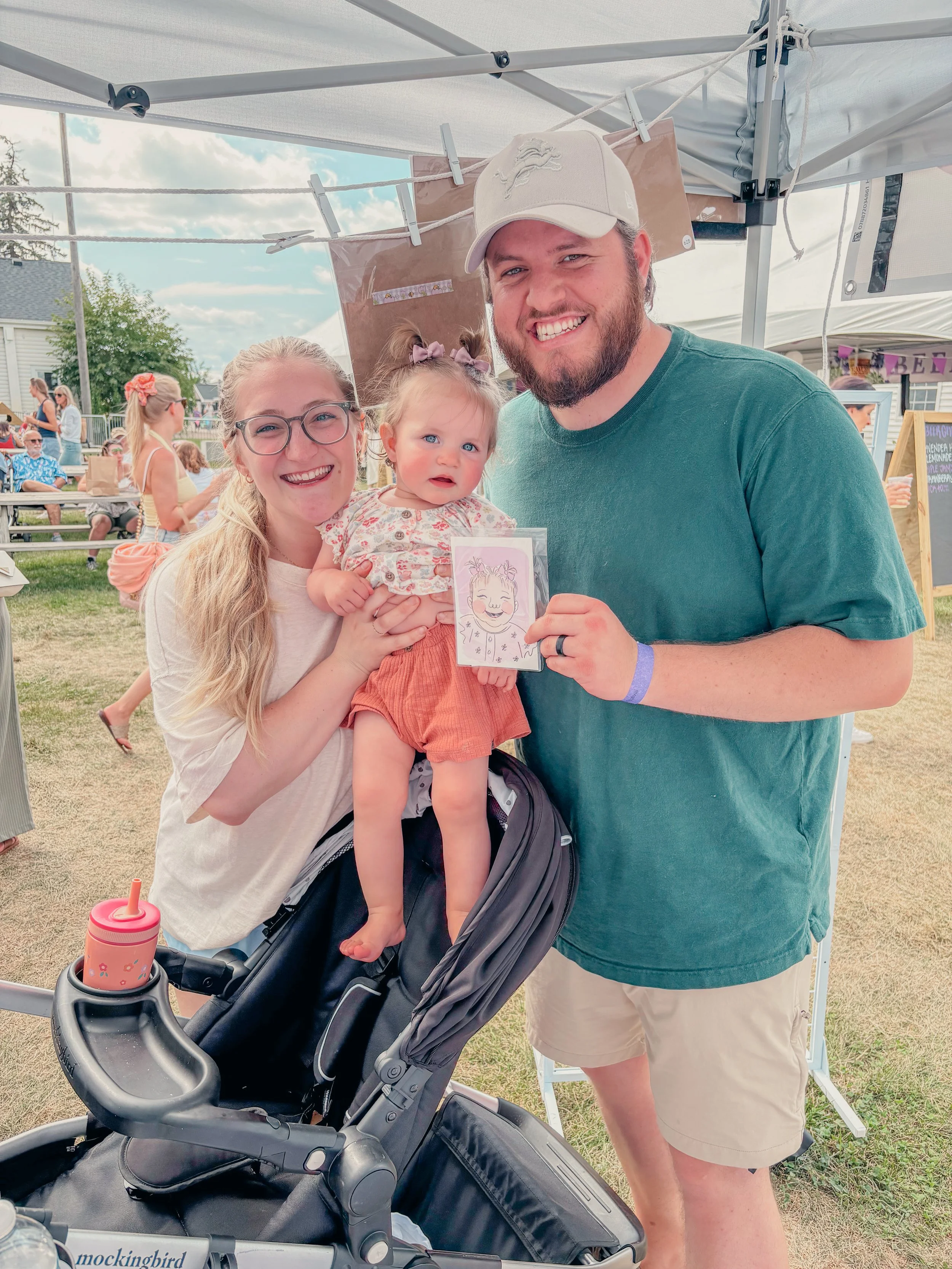 A smiling family outdoors at a fair or festival. A woman holds a little girl with blue eyes and pigtails, and a man stands next to them holding a drawing. The woman and girl are smiling, and the man is also smiling. There is a stroller with a pink cup holder in the foreground.