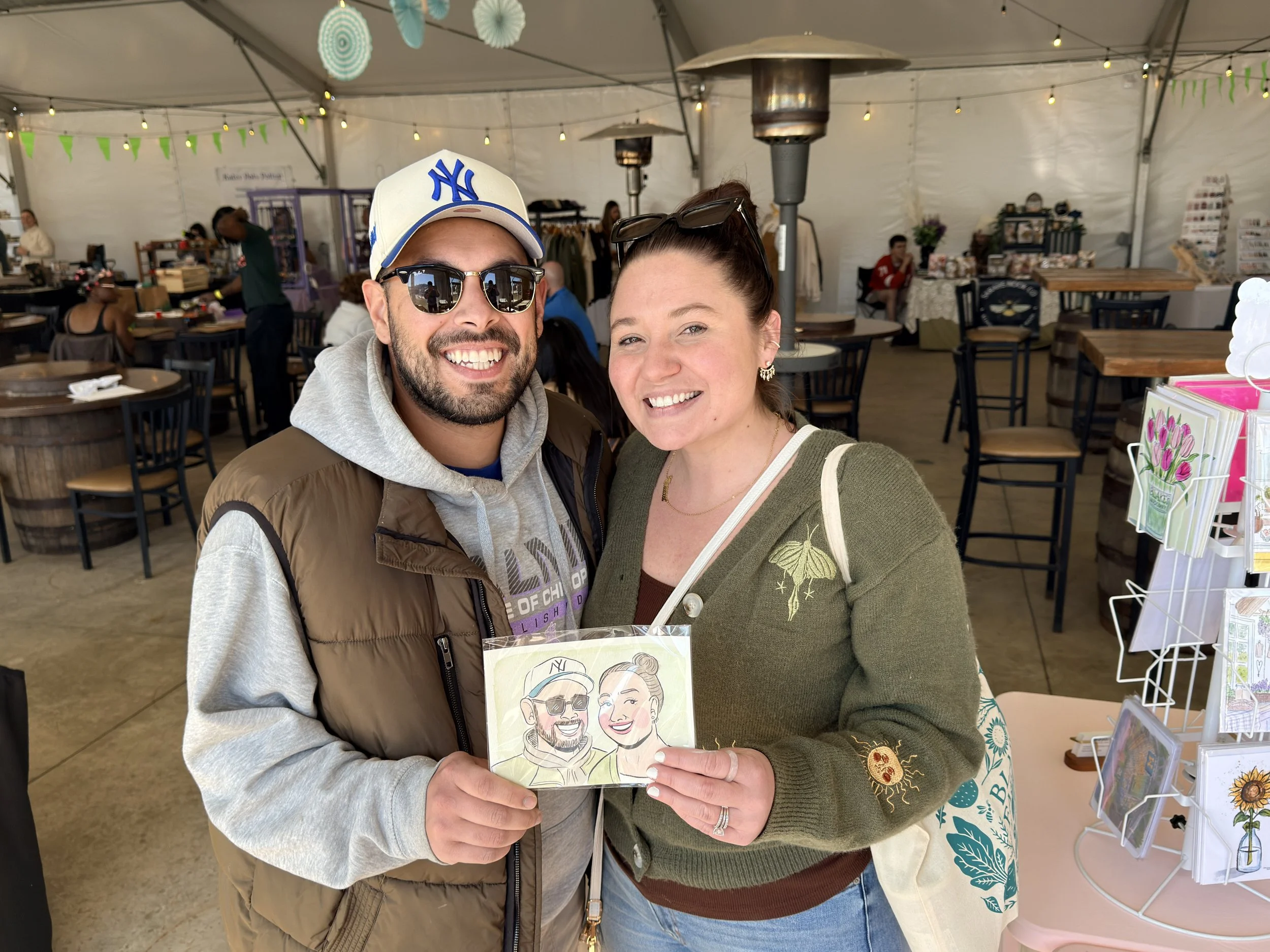 A smiling man in sunglasses, NY Yankees cap, and a brown vest standing next to a smiling woman in sunglasses and a green sweater, holding a caricature drawing of themselves. Inside a tent at a market or fair, with various tables, chairs, and decorative paper lanterns visible.