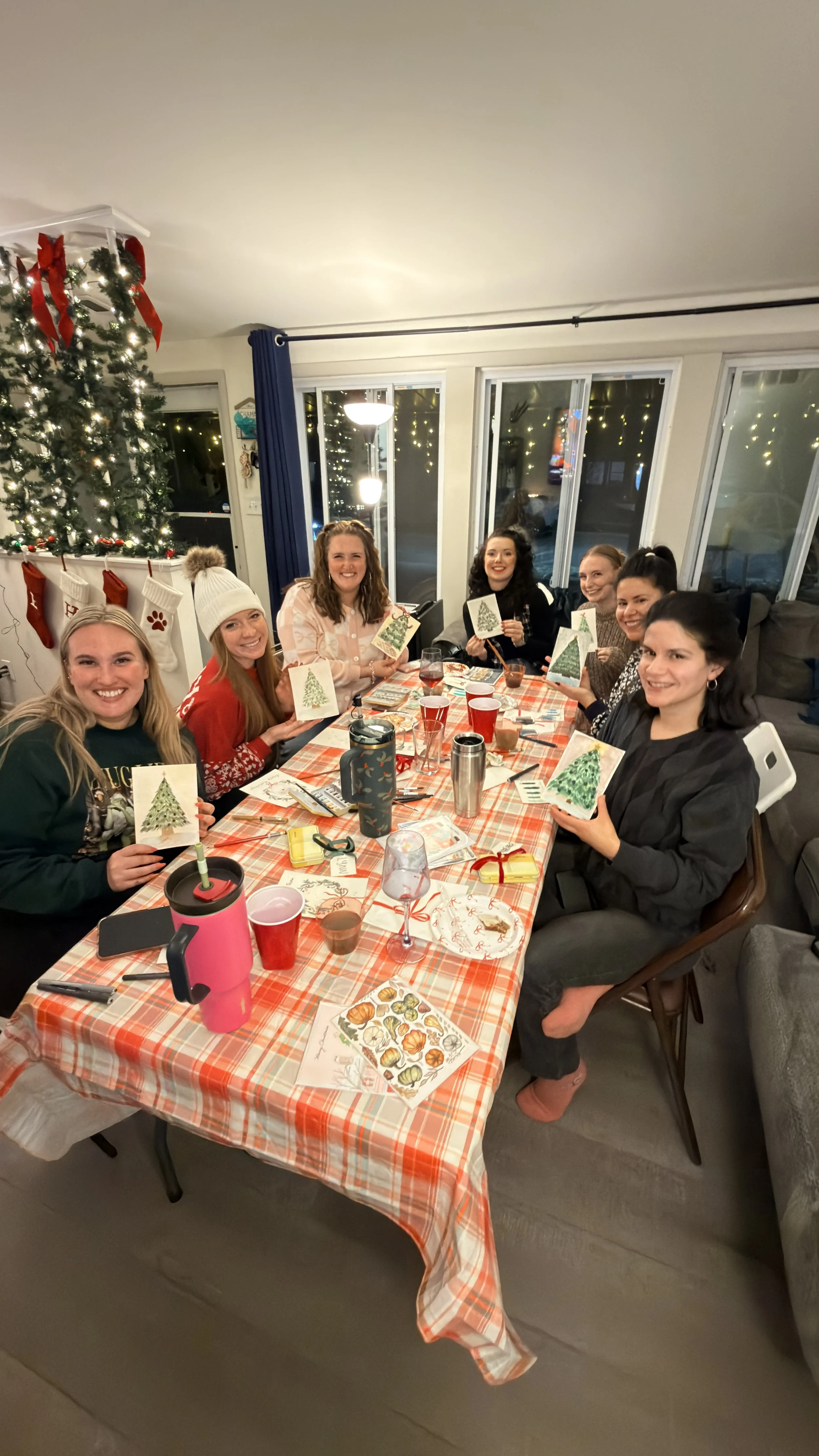 Group of women sitting around a festive table holding Christmas cards, with a decorated Christmas tree in the background.