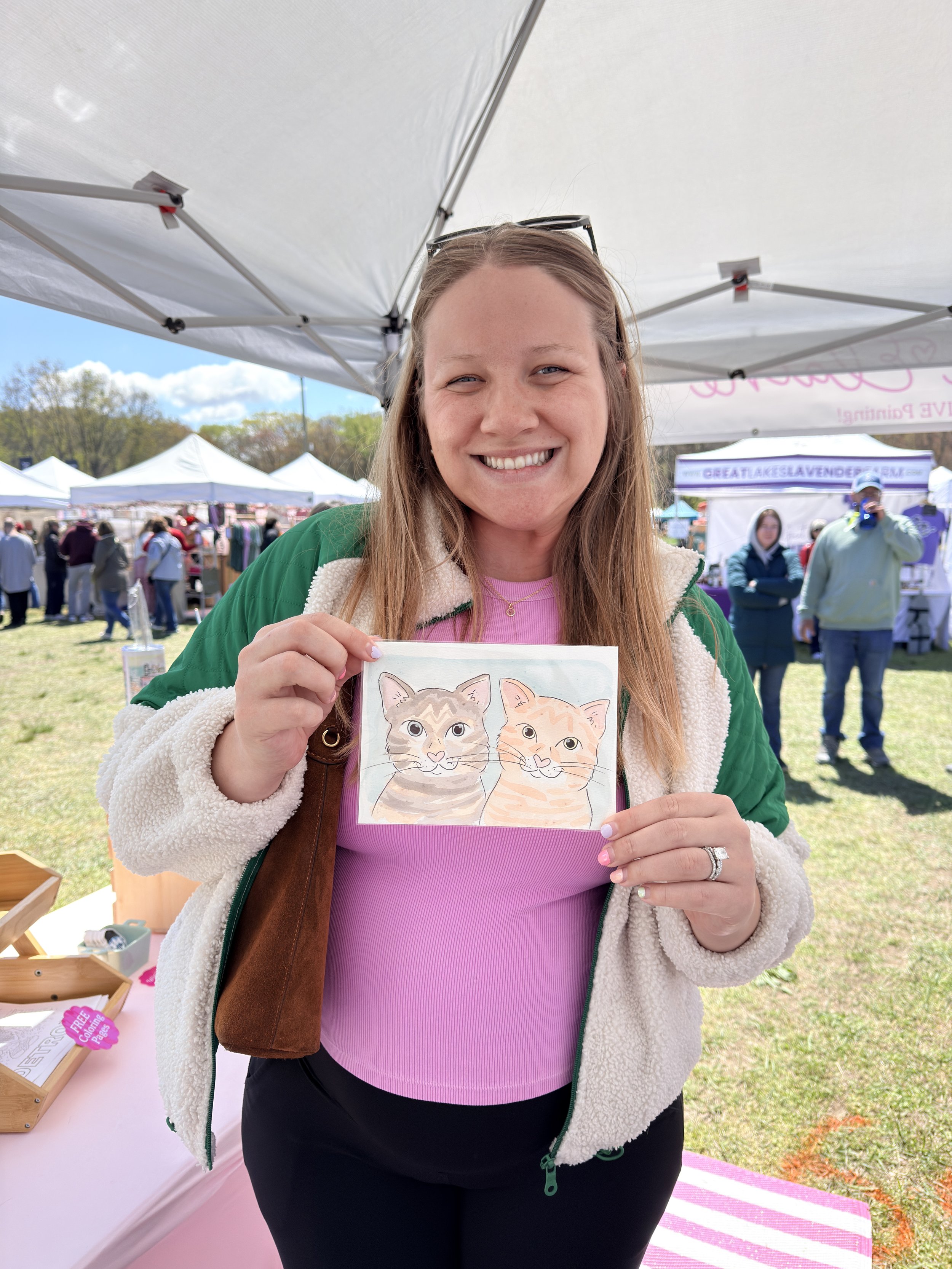 Woman smiling outdoors at a fair or market, holding a colorful drawing of two cats. She wears a green jacket, pink top, and has glasses on her head.