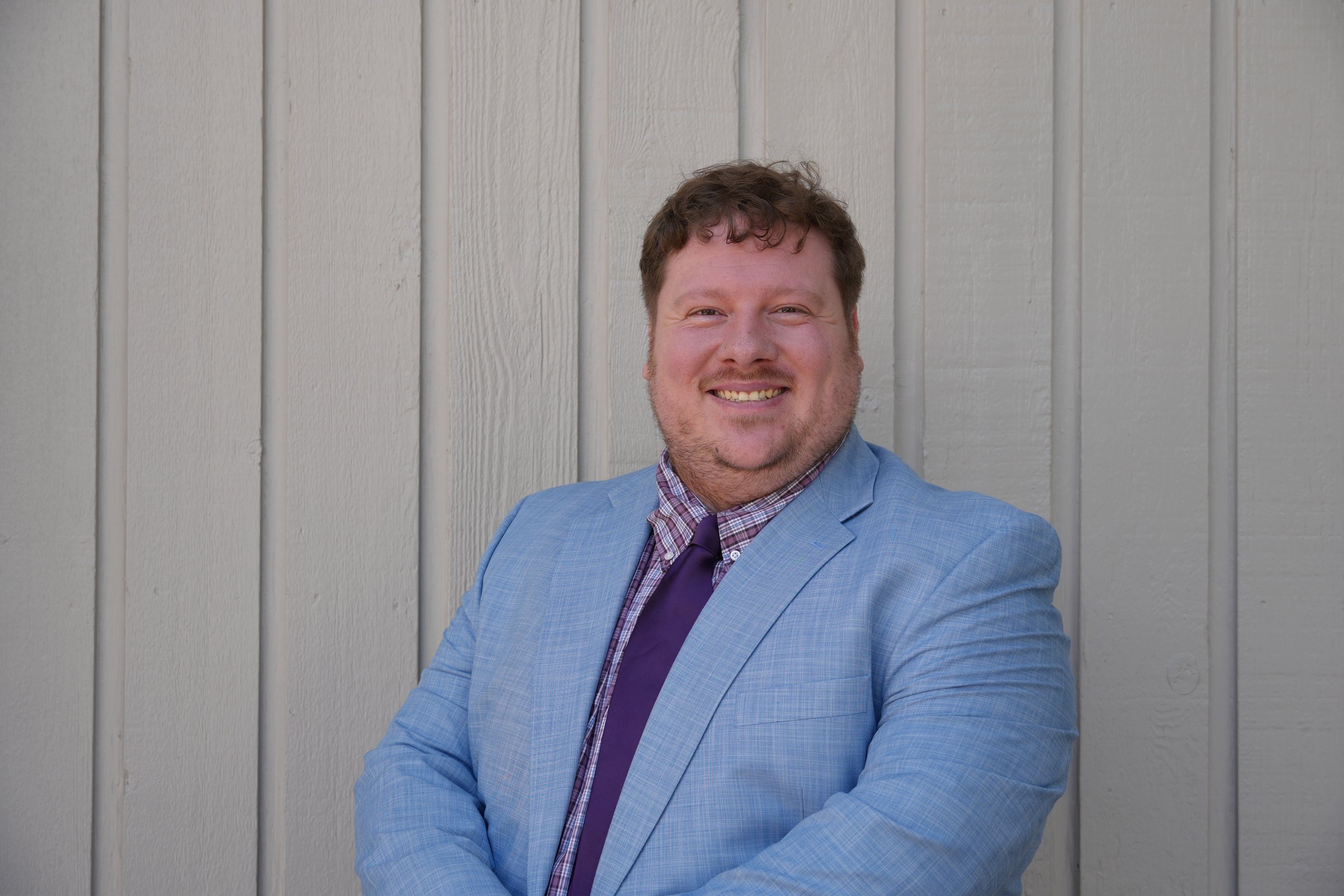 A man in a light blue grey suit, purple checkered shirt, and purple tie smiling with arms crossed in front of a beige wooden wall.
