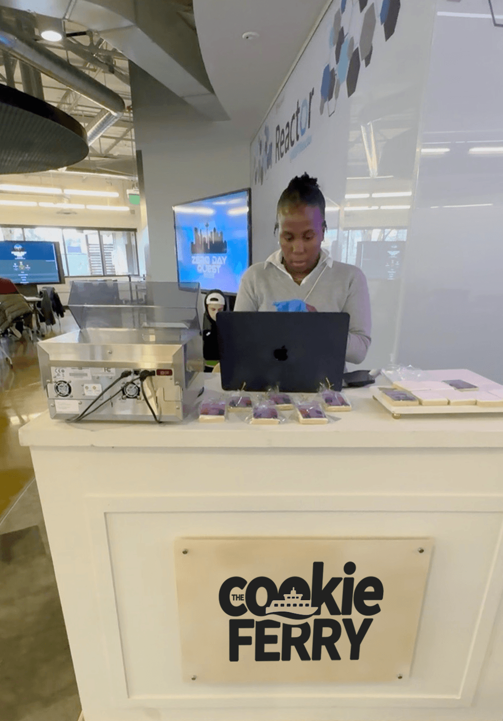 A person working at a kiosk labeled 'The Cookie Ferry' in a modern indoor space, with a laptop and trays of cookies on the counter.