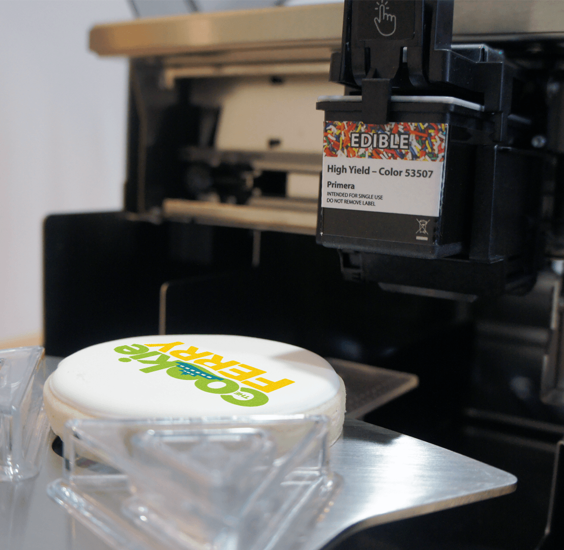 A printed edible cake topper with colorful text that reads 'COOKIES FOR YOU' on a white chocolate surface, with a color label reading 'High Yield - Color 53507 Primera' attached to a black machine in the background.