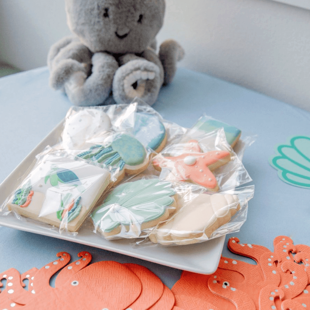A plate of decorated cookies with ocean and beach themes, wrapped in plastic, along with orange octopus-shaped paper decorations, and a plush octopus toy in the background on a blue surface.