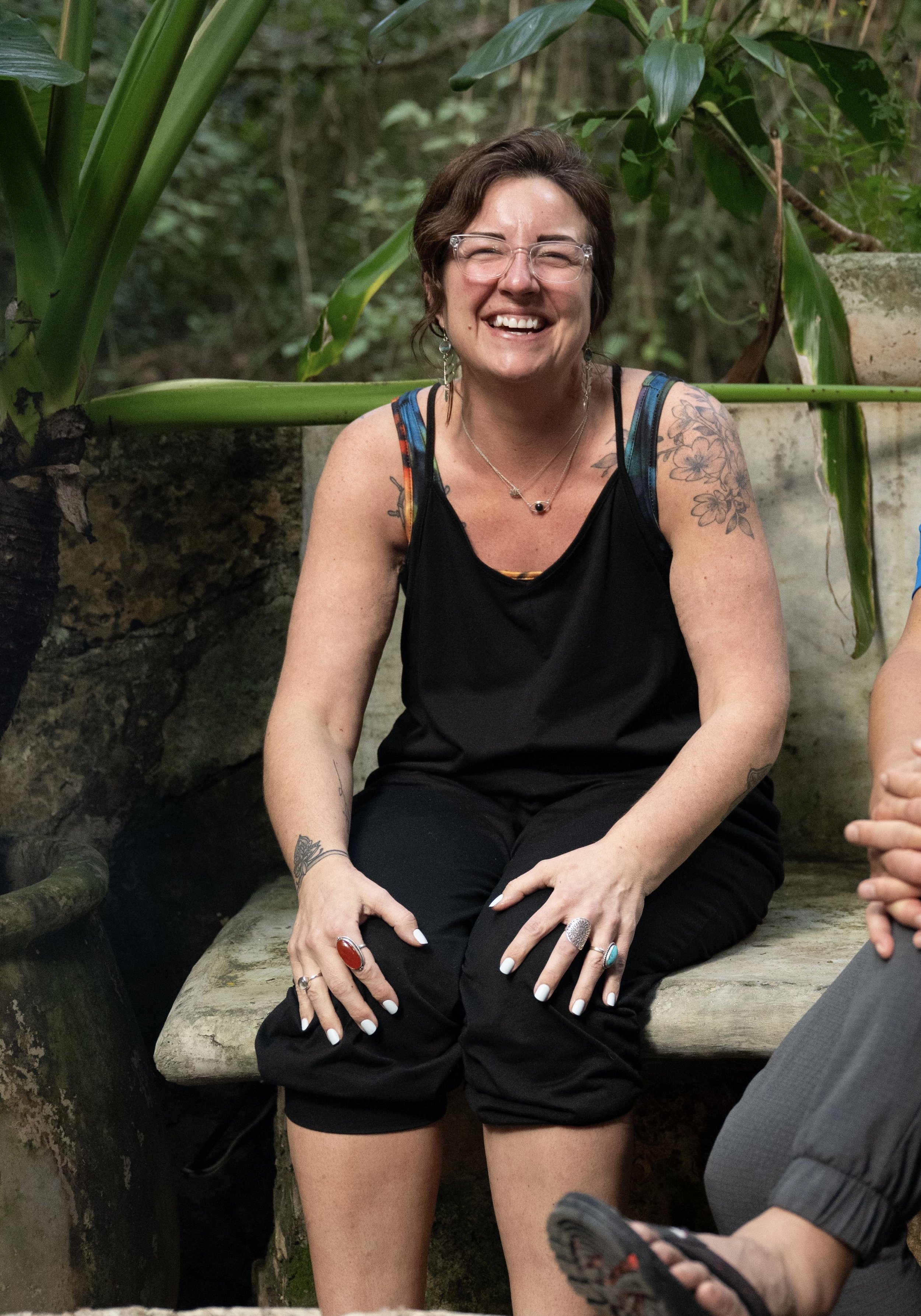 A woman with short brown hair, glasses, and tattoos on her arms, smiling and sitting outdoors surrounded by green foliage.