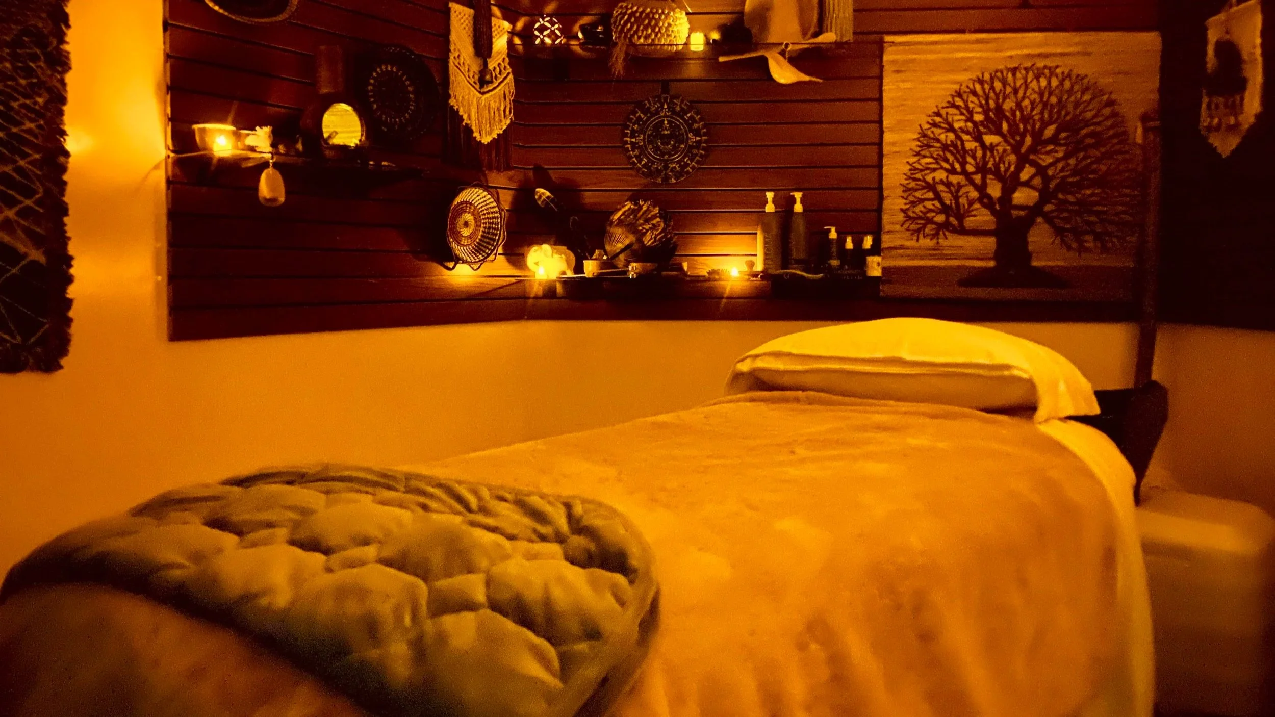 A bedroom with warm lighting featuring a bed with a tufted headboard, a wooden wall décor, candles, bottles, and various decorative items including a large artwork of a tree on the wall.