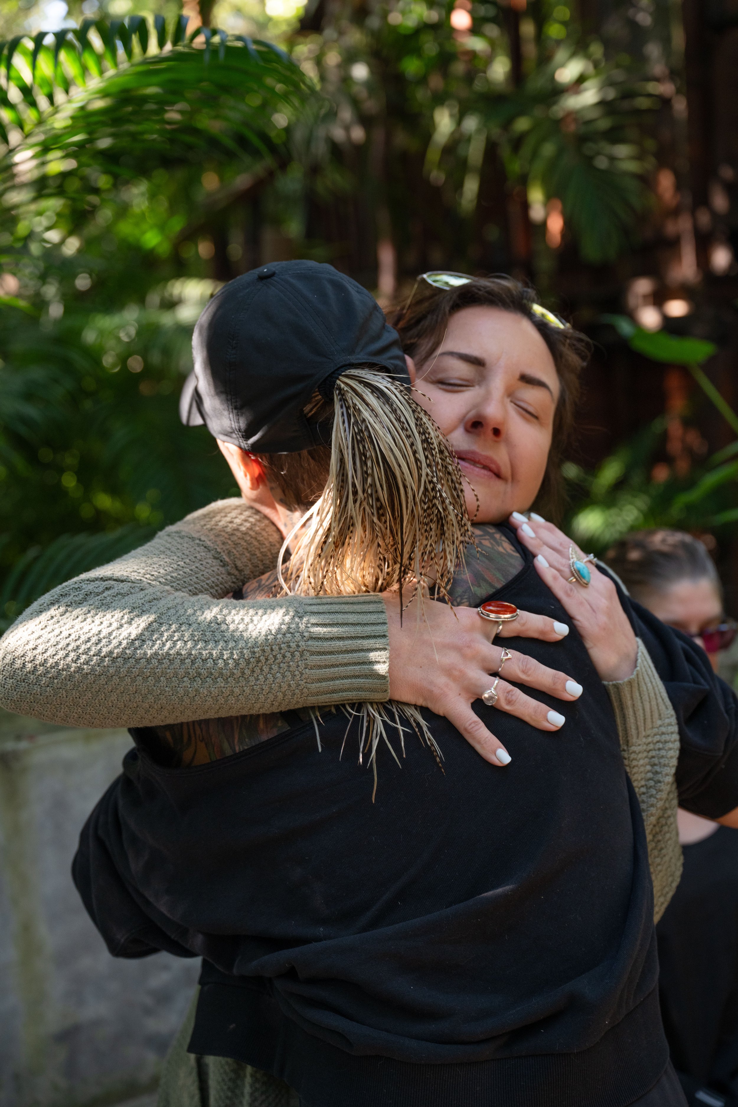 Two women are sharing a warm hug outdoors surrounded by green foliage, with one woman smiling and the other partially visible.