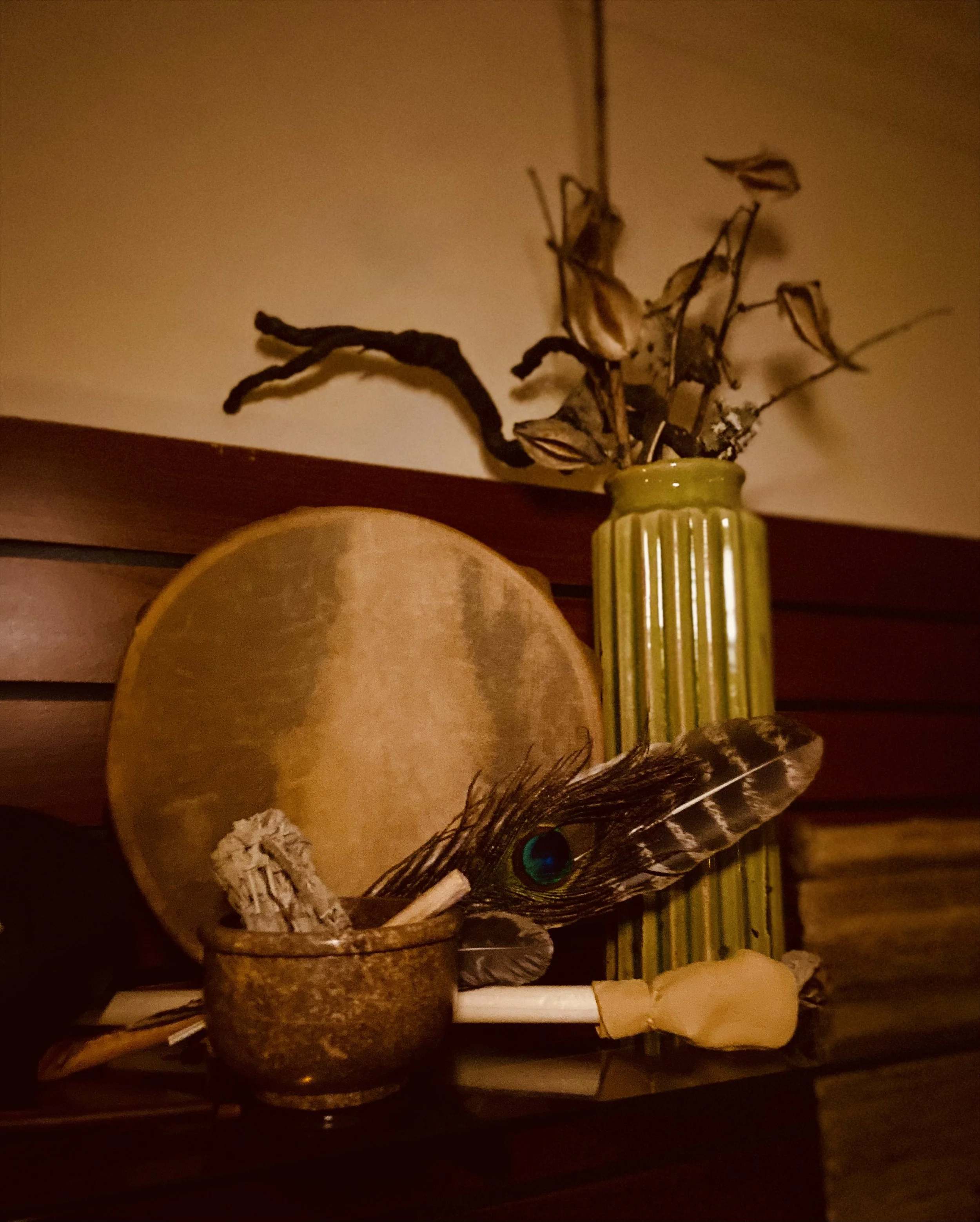 A decorative display includes a brown pitcher with a peacock feather and a white candle, in front of a large round object, possibly a clock or mirror, on a dark wood shelf.