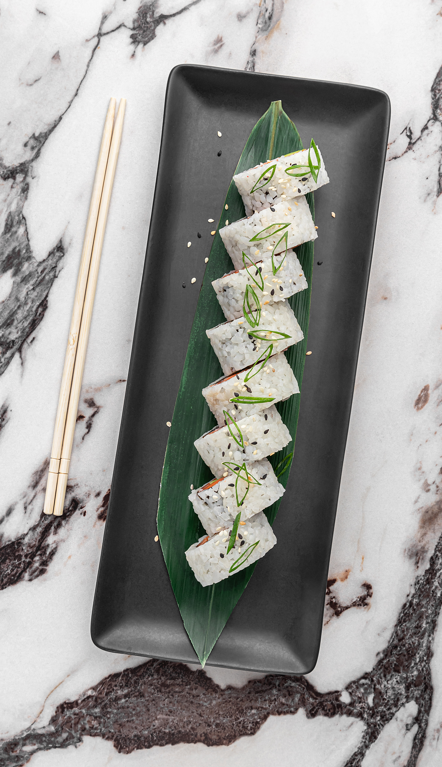 Six pieces of sushi arranged on a green leaf, garnished with sliced green onions, on a black rectangular plate with chopsticks to the side.