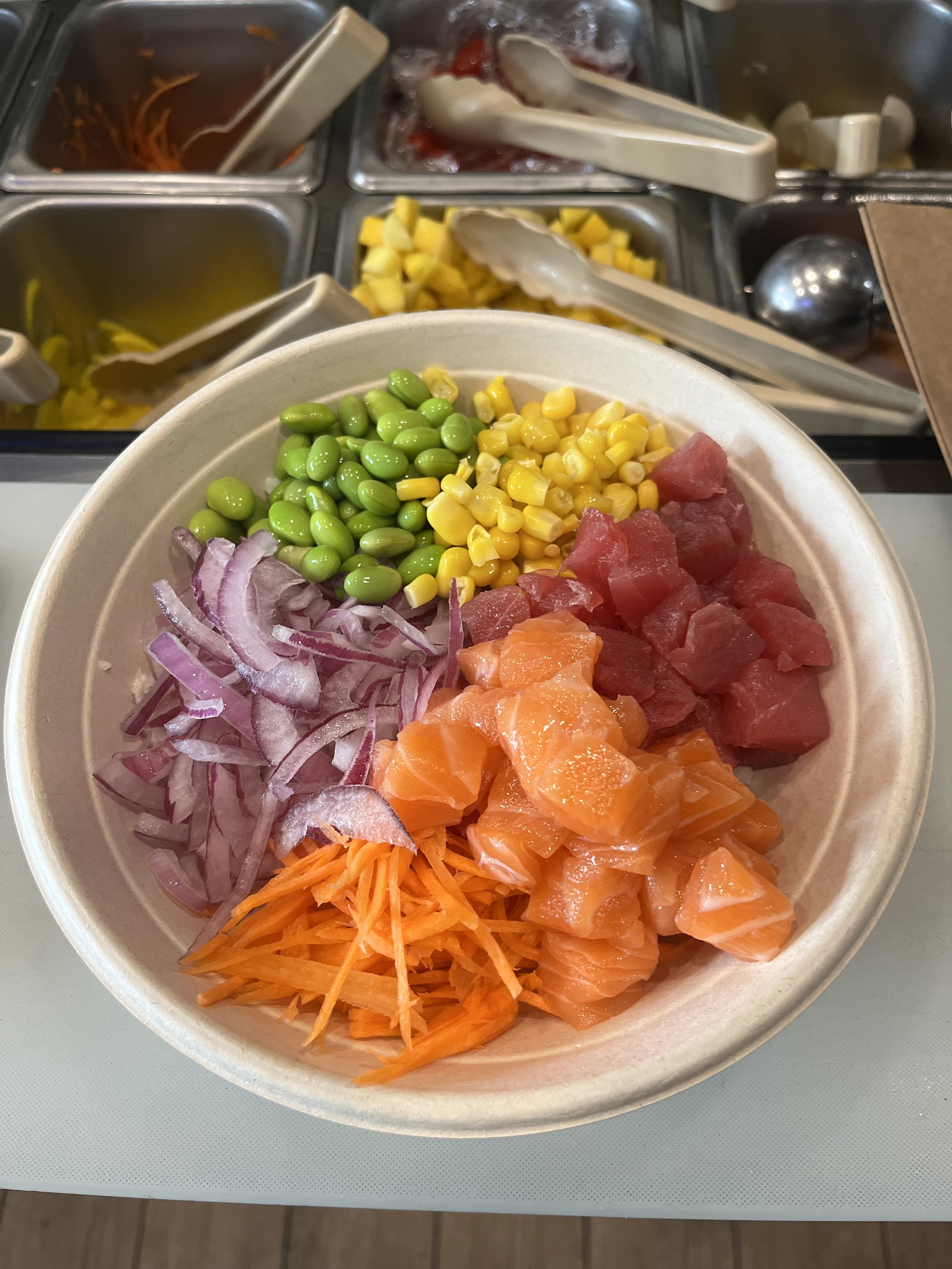 A bowl with sliced red onions, shredded carrots, diced green edamame, corn, and salmon, with a buffet in the background.